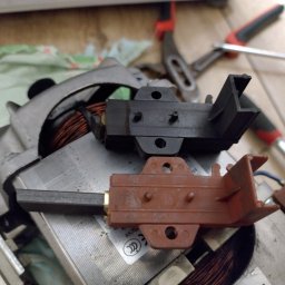 KK SERWIS KAMIL KOCYŁA - Szczotki silnika pralki, czarne i brązowe, leżące na silniku z miedzianymi uzwojeniami, obok narzędzi na tle roboczym. Detal z bliska.