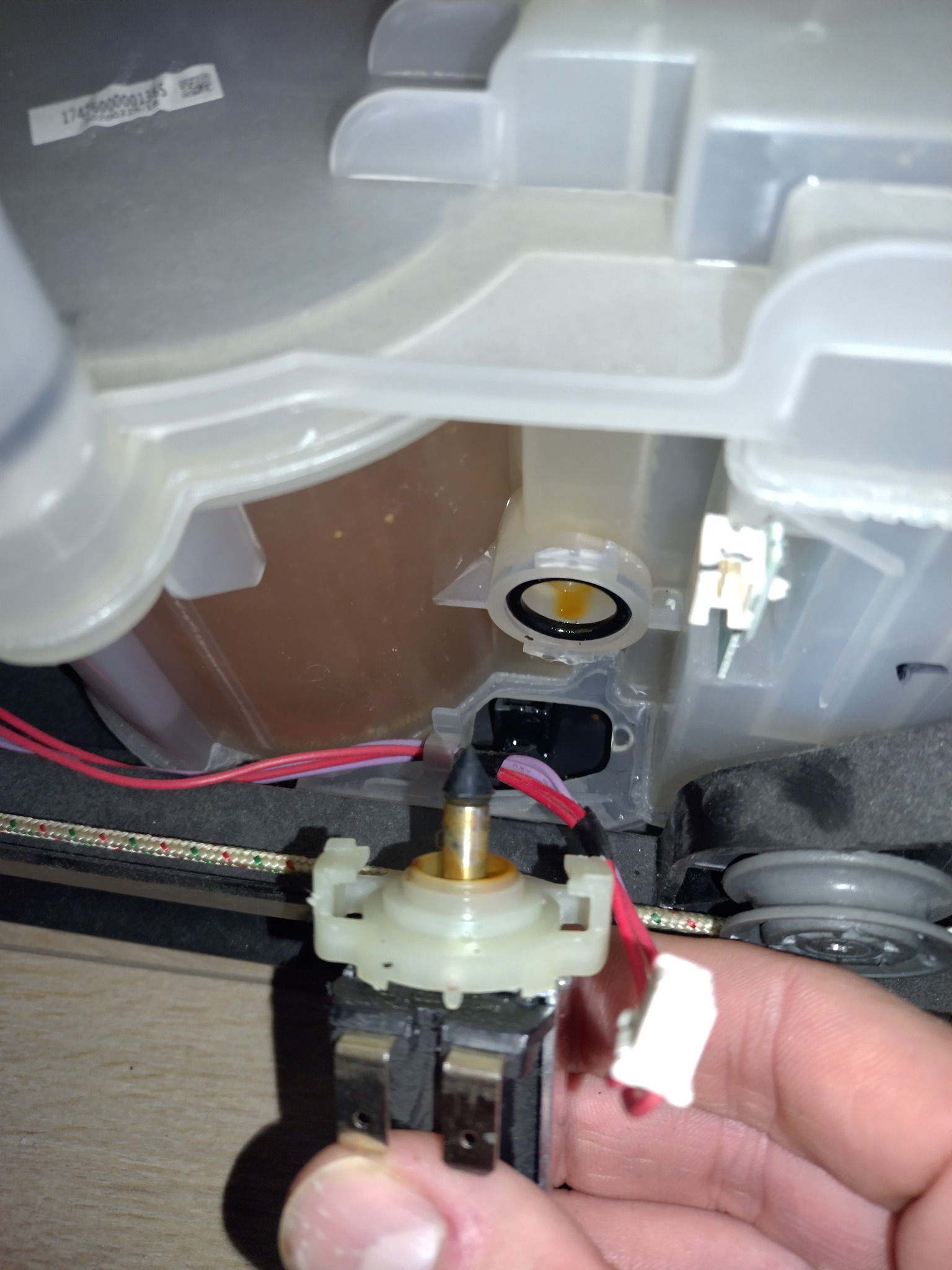 Zbliżenie na element zmywarki w trakcie naprawy: widoczny czujnik poziomu wody, przewody i dłoń serwisanta. W tle zbiornik z brudną wodą. Detal naprawy AGD.