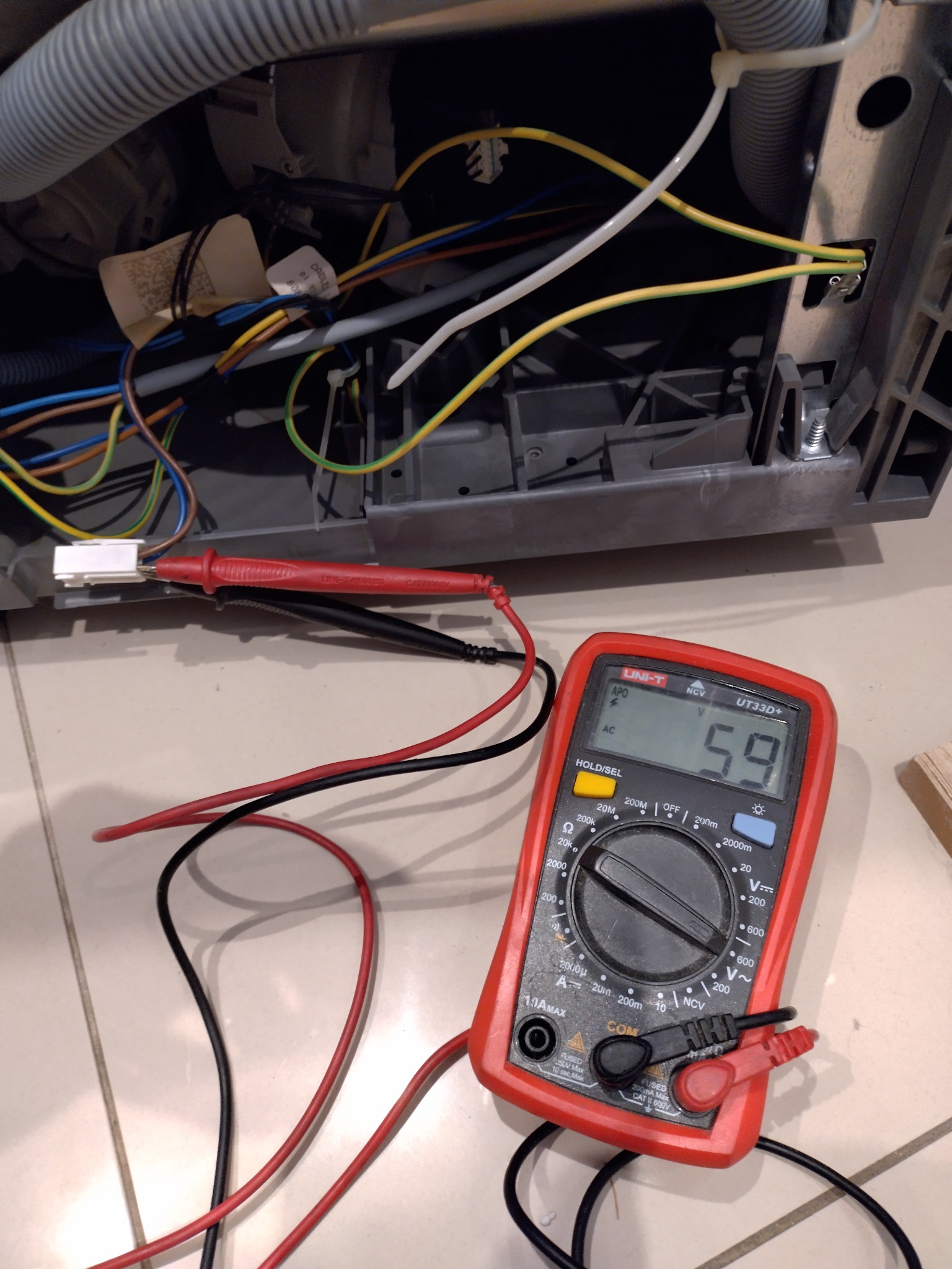 Multimetr UNI-T UT33D+ wskazuje 59 V przy przewodach z tyłu szarej obudowy urządzenia AGD. Widoczne kable, złącza i elementy konstrukcyjne.