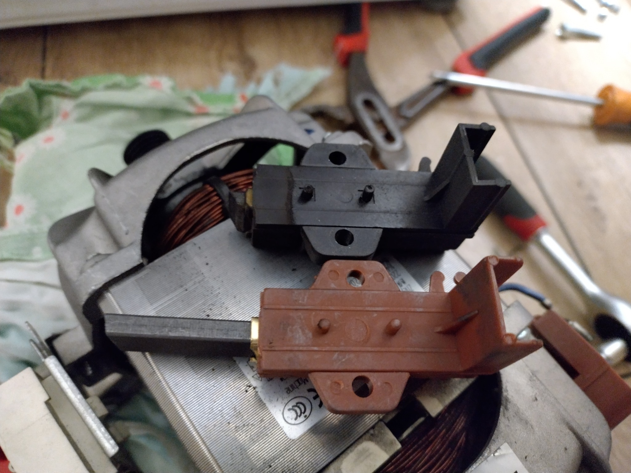 Szczotki silnika pralki, czarne i brązowe, leżące na silniku z miedzianymi uzwojeniami, obok narzędzi na tle roboczym. Detal z bliska.