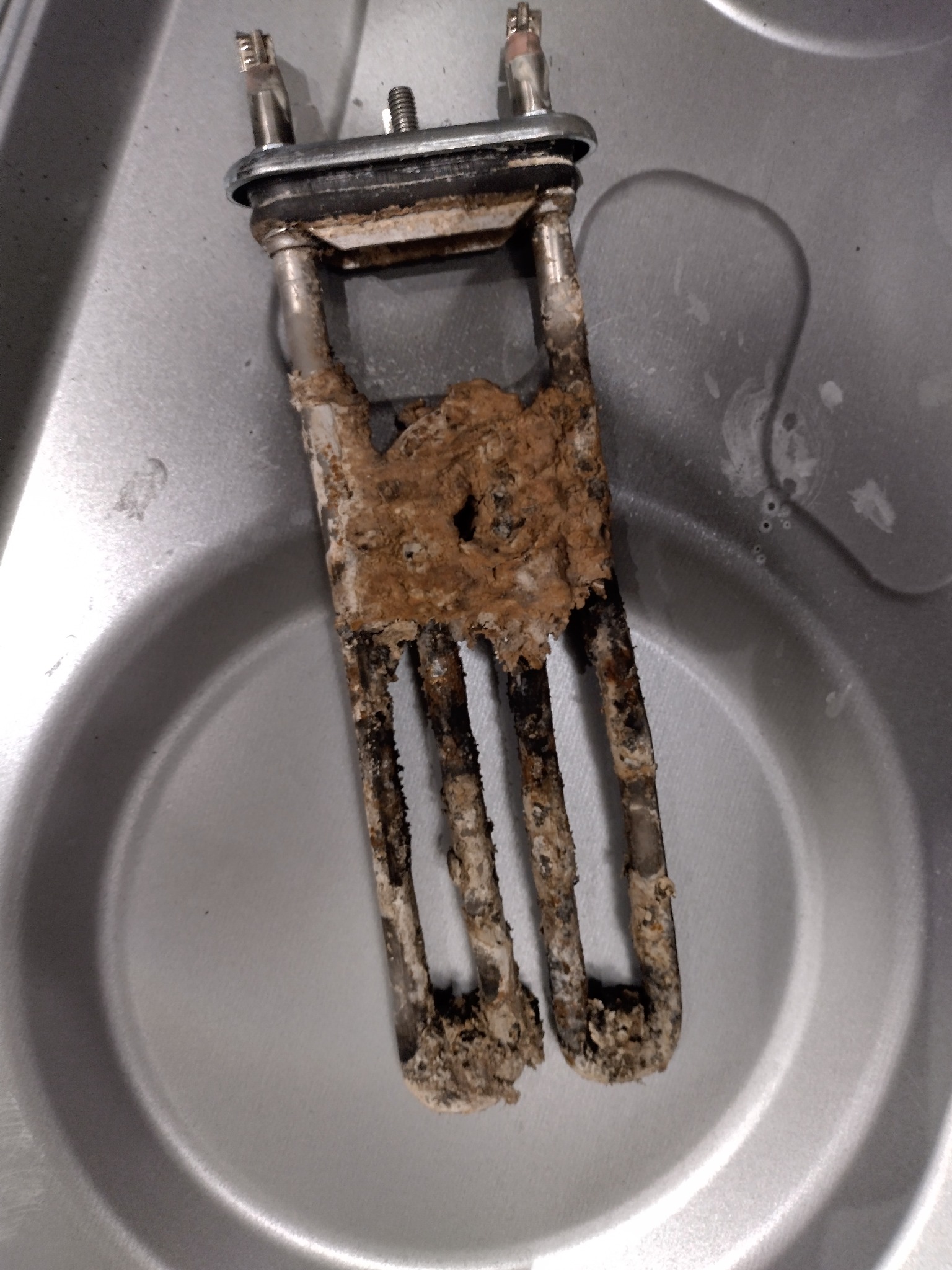 Zdemontowana, silnie zakamieniona grzałka z pralki, widoczne osady wapienne i korozja, leżąca w metalowej misce. Detal z bliska, ukazujący stopień zużycia elementu.