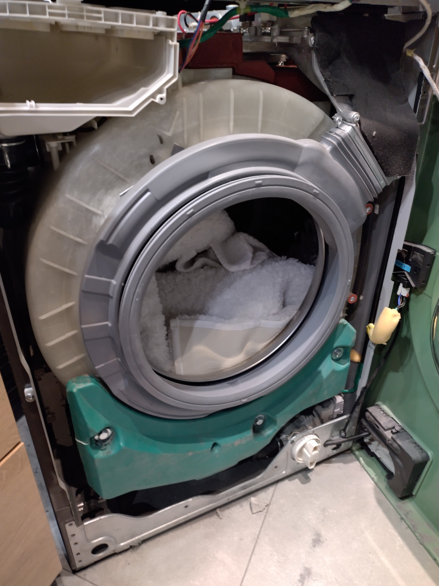 Otwarta pralka z widocznym bębnem wypełnionym białymi tkaninami. Widoczne elementy konstrukcyjne i okablowanie. Ujęcie z bliska, ukazujące wnętrze urządzenia.
