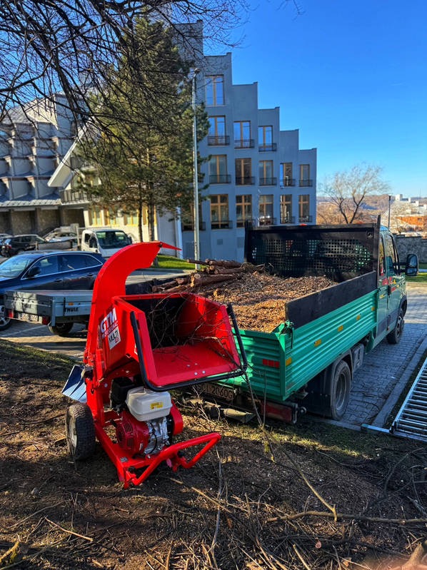 Czerwony rębak do gałęzi GTM Multi 40 na tle zielonej ciężarówki z ładunkiem rozdrobnionych gałęzi, budynki w tle, słoneczny dzień.