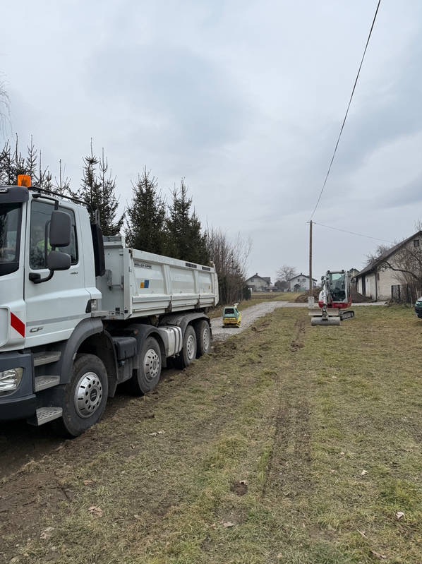 Ciężarówka i minikoparka na działce, przygotowanie terenu pod budowę. Widoczne ślady opon na ziemi. Pochmurne niebo w tle.