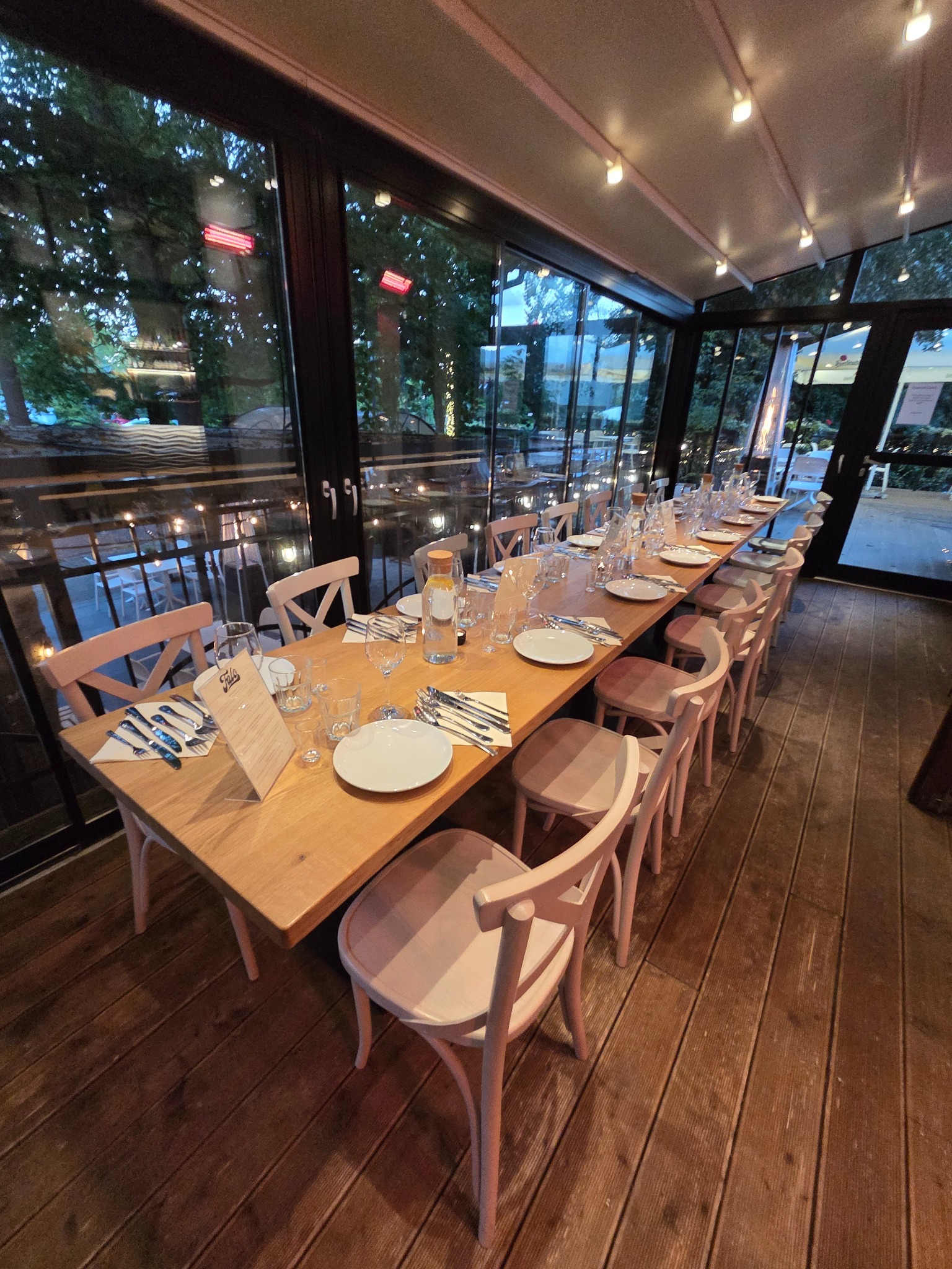 Długi stół z jasnego drewna, nakryty zastawą dla gości, otoczony krzesłami w odcieniach różu. Widok przez przeszkloną ścianę na zewnątrz, oświetlenie sufitowe.