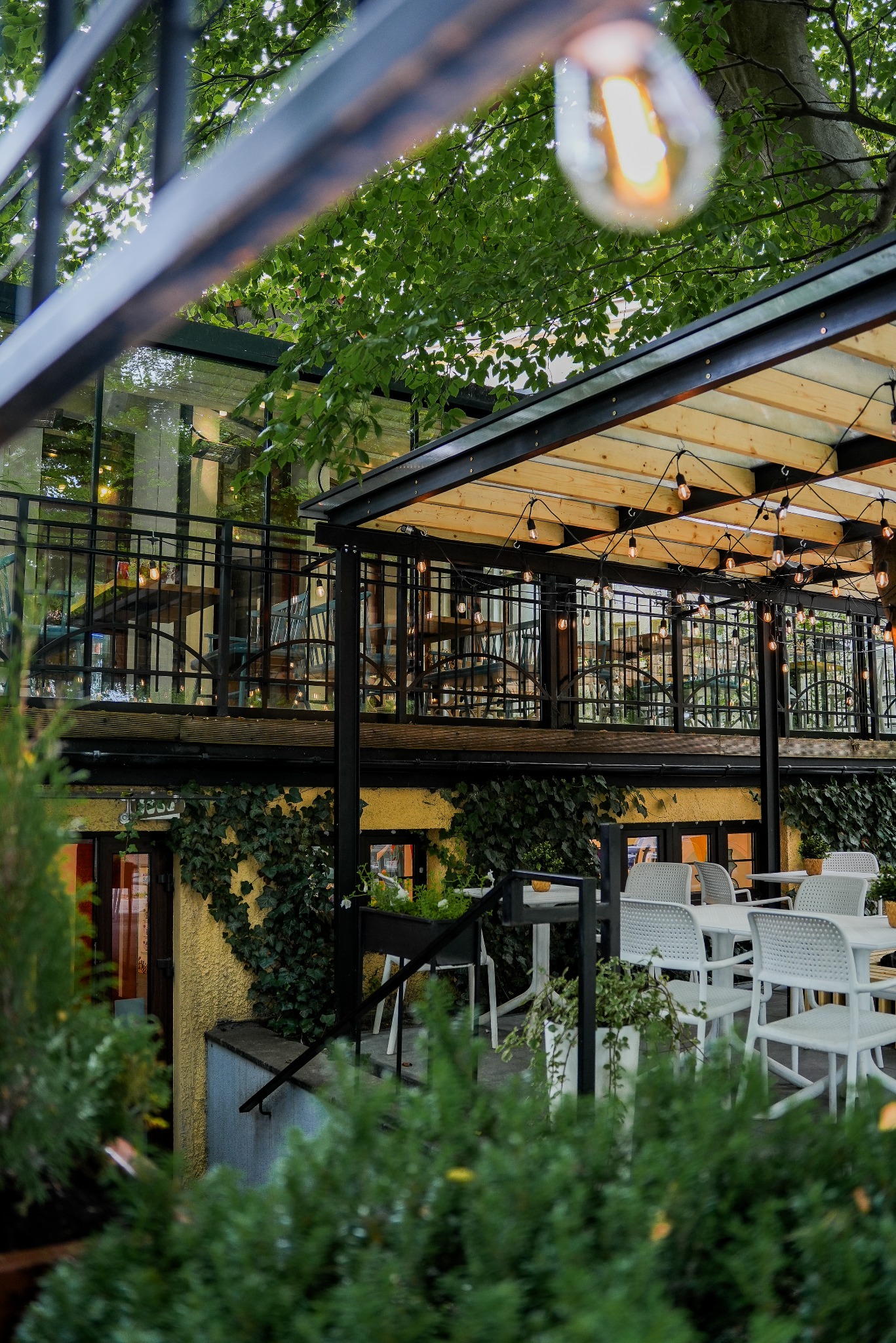 Urokliwa restauracja z tarasem, oświetlona girlandami żarówek, otoczona zielenią. Widok z dołu, podkreślający atmosferę miejsca.