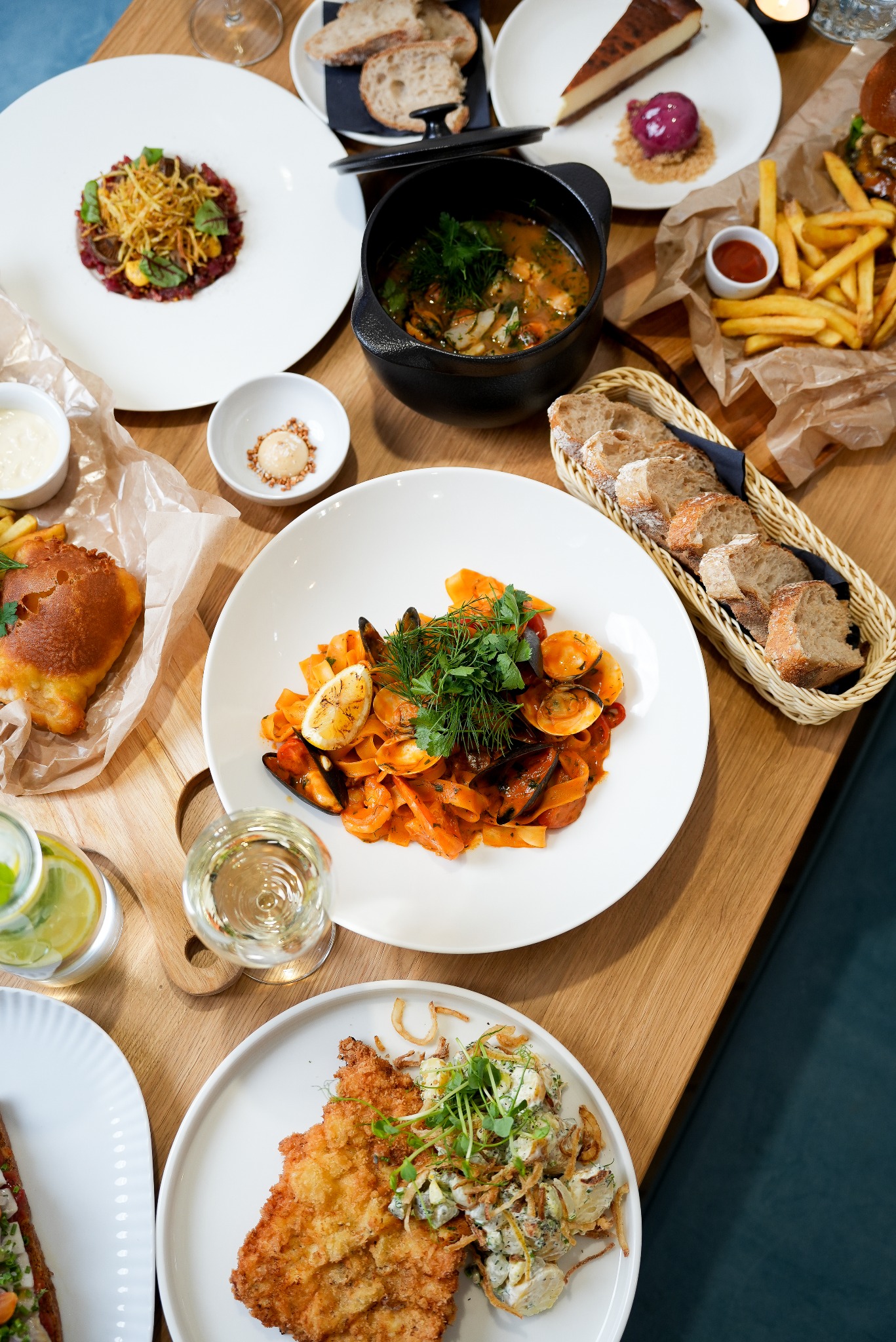 Stół z różnorodnymi daniami: makaron z owocami morza, zupa rybna w czarnym garnku, tatar wołowy, sznycel, frytki, pieczywo i deser. Jasne drewno blatu, biała zastawa.