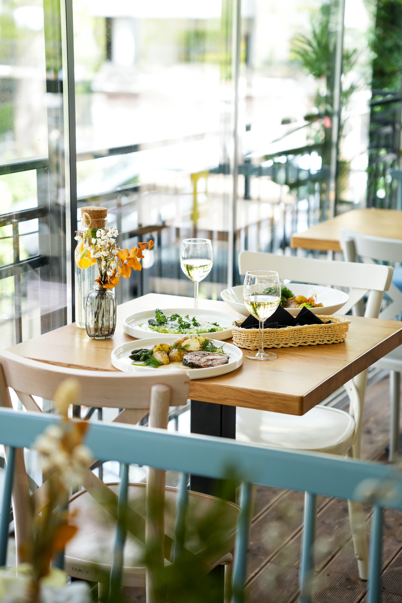 Nakryty stół z daniami i winem w restauracji z widokiem na zieleń. Dekoracja kwiatowa w szklanym wazonie. Jasne, naturalne światło.
