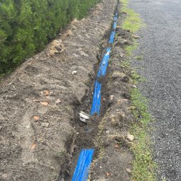 Mariusz Kusiak - Wykop z niebieskimi rurami kablowymi ułożonymi w ziemi, widok z góry, obok ścieżka z kamieni. Etap prac przyłącza elektrycznego.