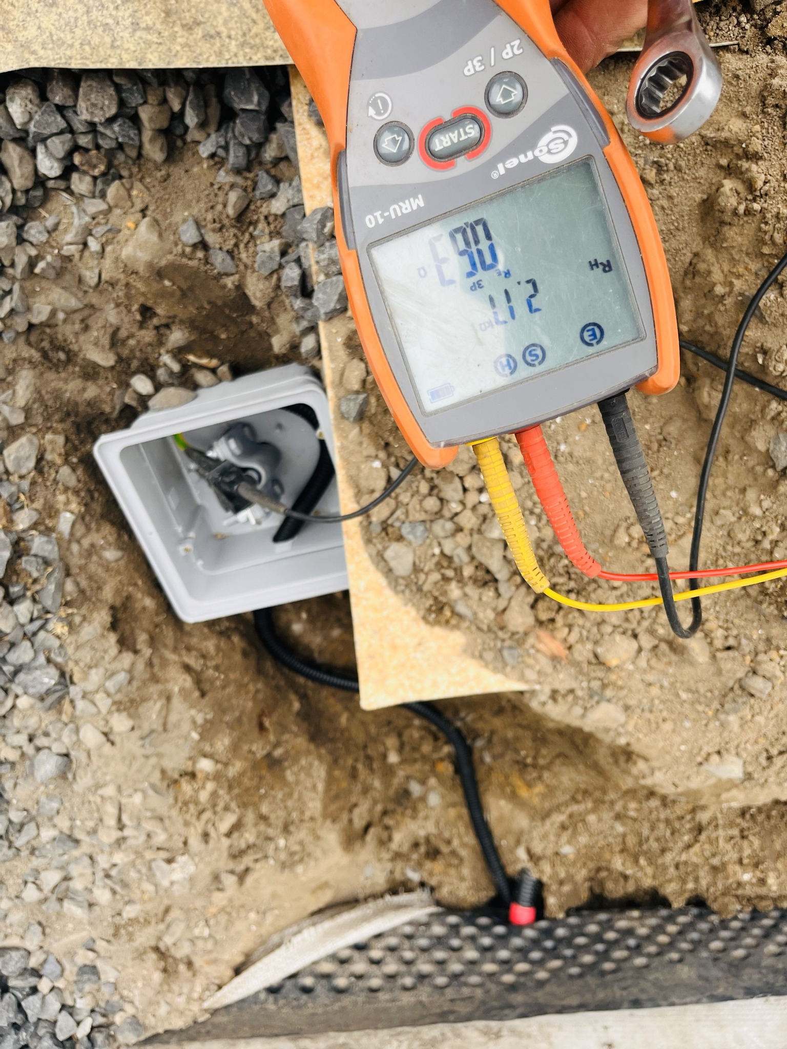 Pomiar uziemienia: Miernik Sonel MRU-10 z przewodami podłączony do skrzynki elektrycznej w wykopie, obok leży klucz. Widoczne kamienie i ziemia.