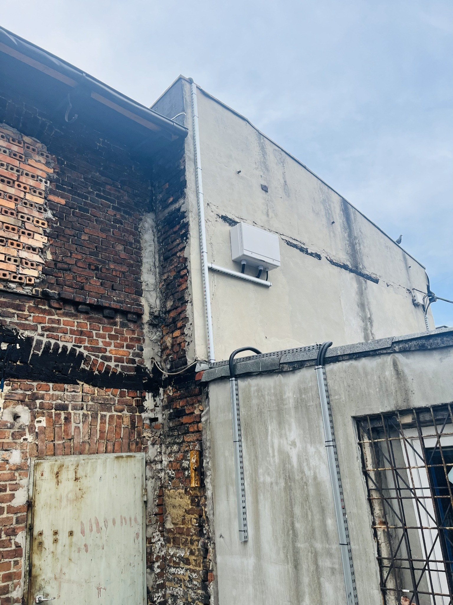 Biała skrzynka elektryczna zamontowana na elewacji budynku z widocznymi przewodami i elementami instalacji. Widok z dołu na budynek z cegły i betonu.