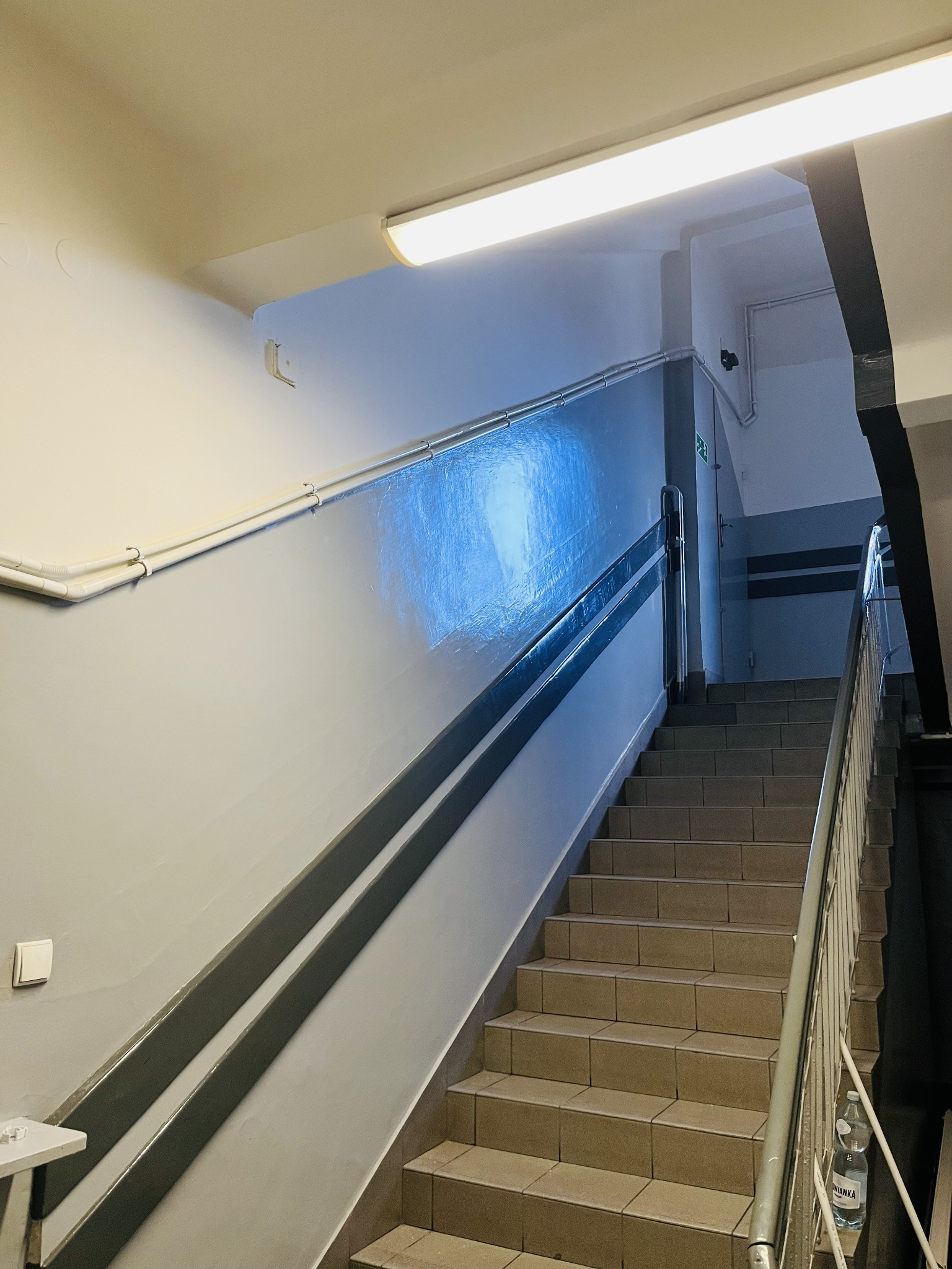 Klatka schodowa z oświetleniem jarzeniowym i widocznymi przewodami elektrycznymi na ścianie. Szare barierki i białe ściany. Widoczny włącznik światła.