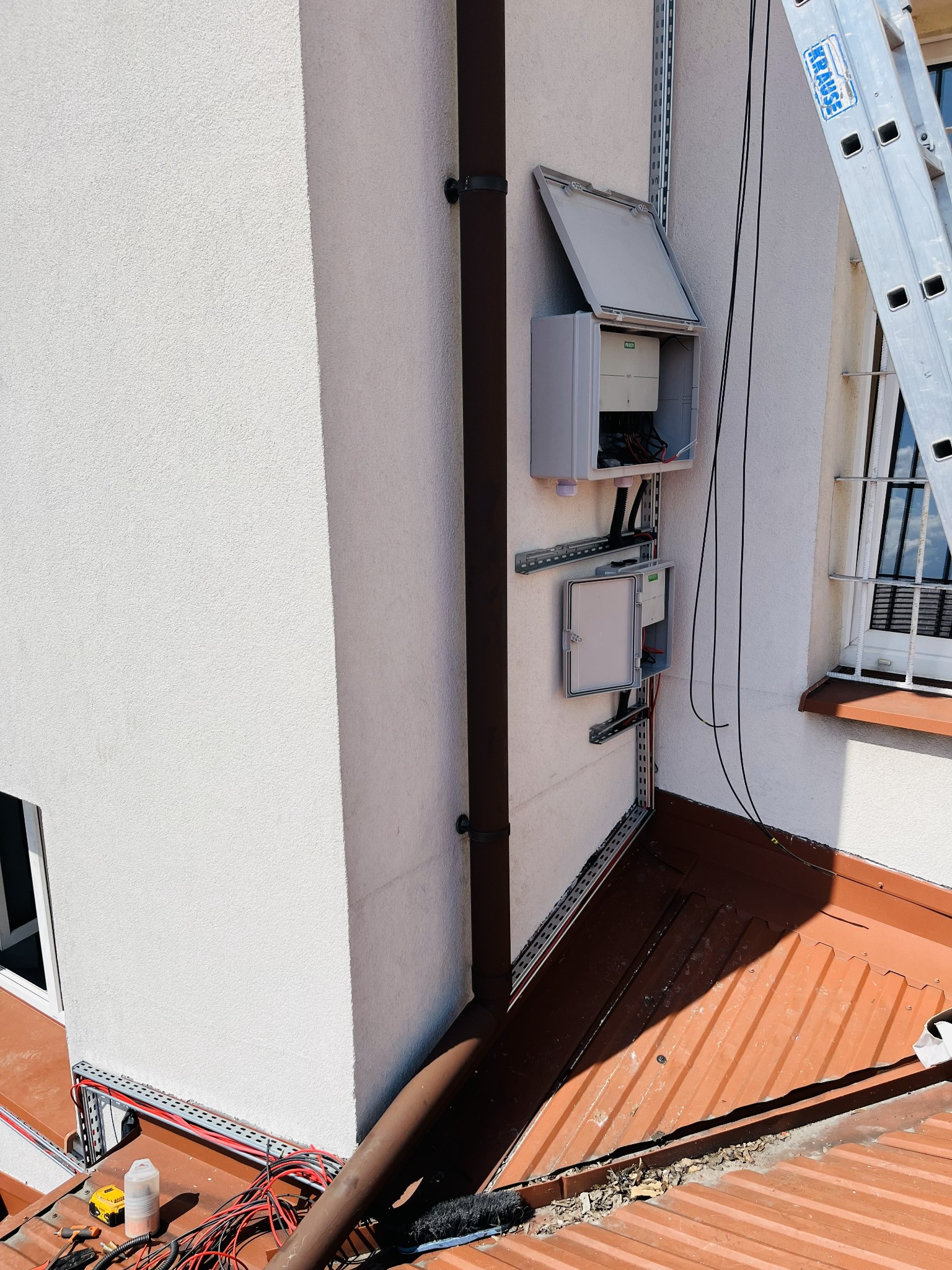 Dwa szare panele elektryczne zamontowane na elewacji budynku, połączone przewodami. Widoczna drabina i narzędzia na dachu. Instalacja w trakcie prac.