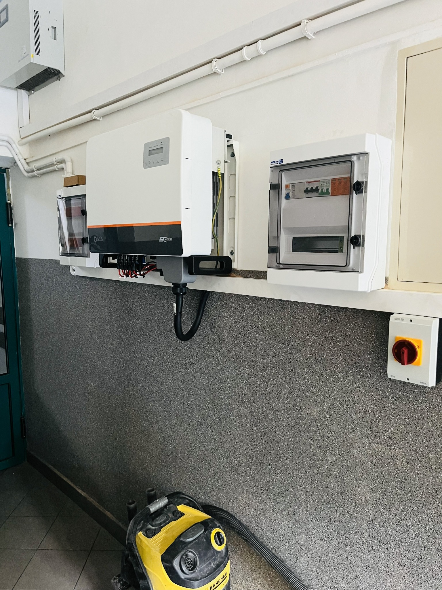 Biała skrzynka elektryczna z bezpiecznikami, inwerter solarny Solis i wyłącznik prądu zamontowane na ścianie. Na podłodze odkurzacz Karcher.