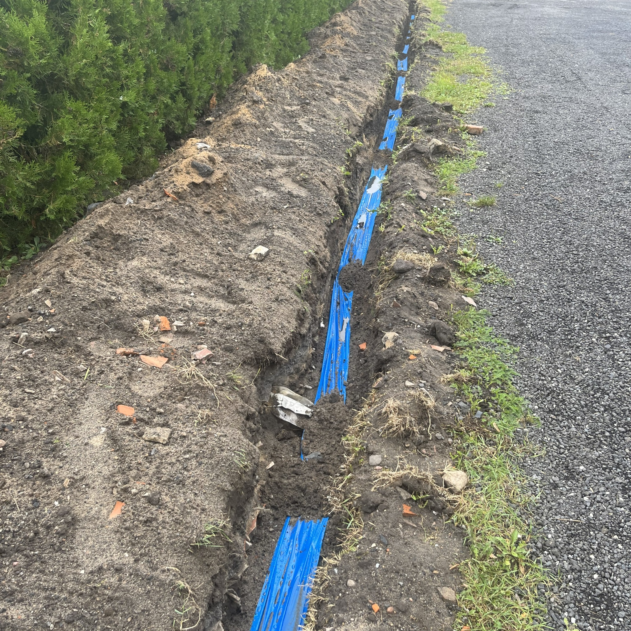 Wykop z niebieskimi rurami kablowymi ułożonymi w ziemi, widok z góry, obok ścieżka z kamieni. Etap prac przyłącza elektrycznego.