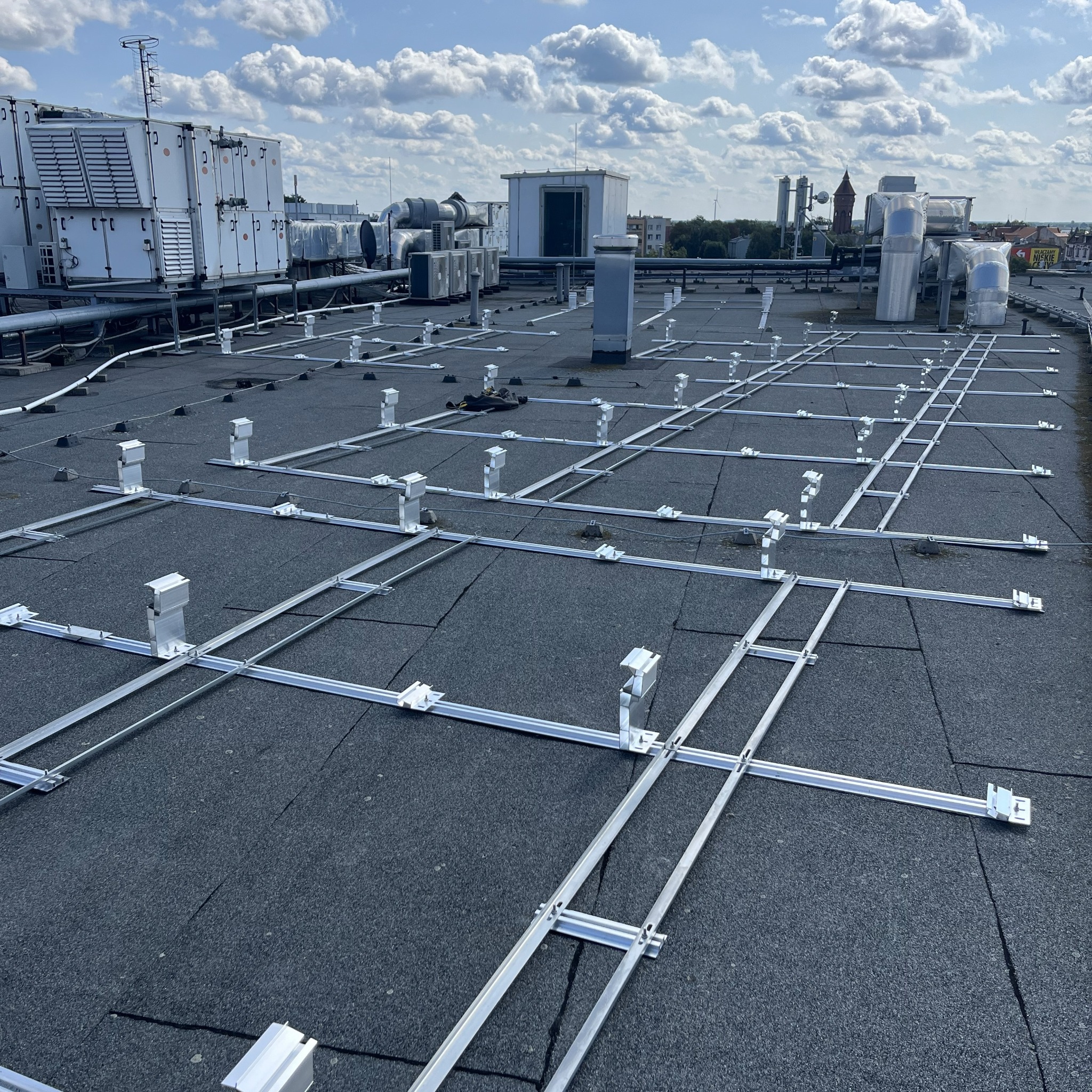Metalowa konstrukcja wsporcza na dachu budynku, prawdopodobnie pod panele fotowoltaiczne. W tle widoczne urządzenia wentylacyjne i budynki miejskie.
