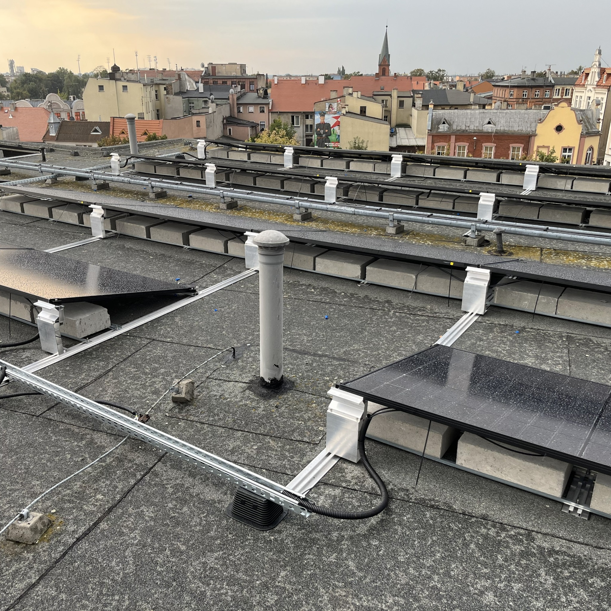 Panele słoneczne na dachu budynku z widokiem na miasto w tle, instalacja w trakcie montażu, widoczne elementy mocujące i przewody.