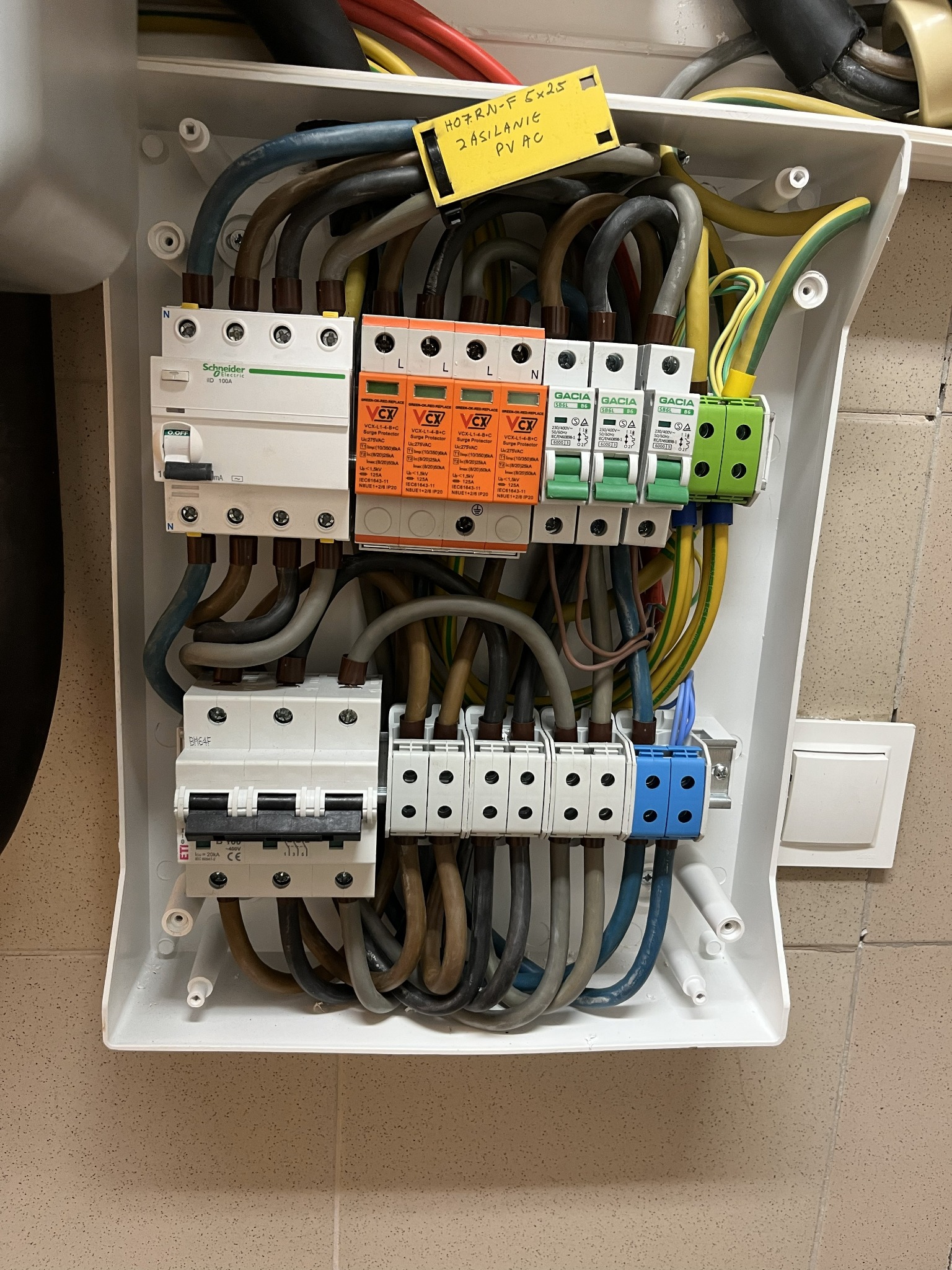 Rozdzielnia elektryczna z zabezpieczeniami przepięciowymi, widoczne kable, bezpieczniki i wyłączniki. Żółta etykieta z opisem.