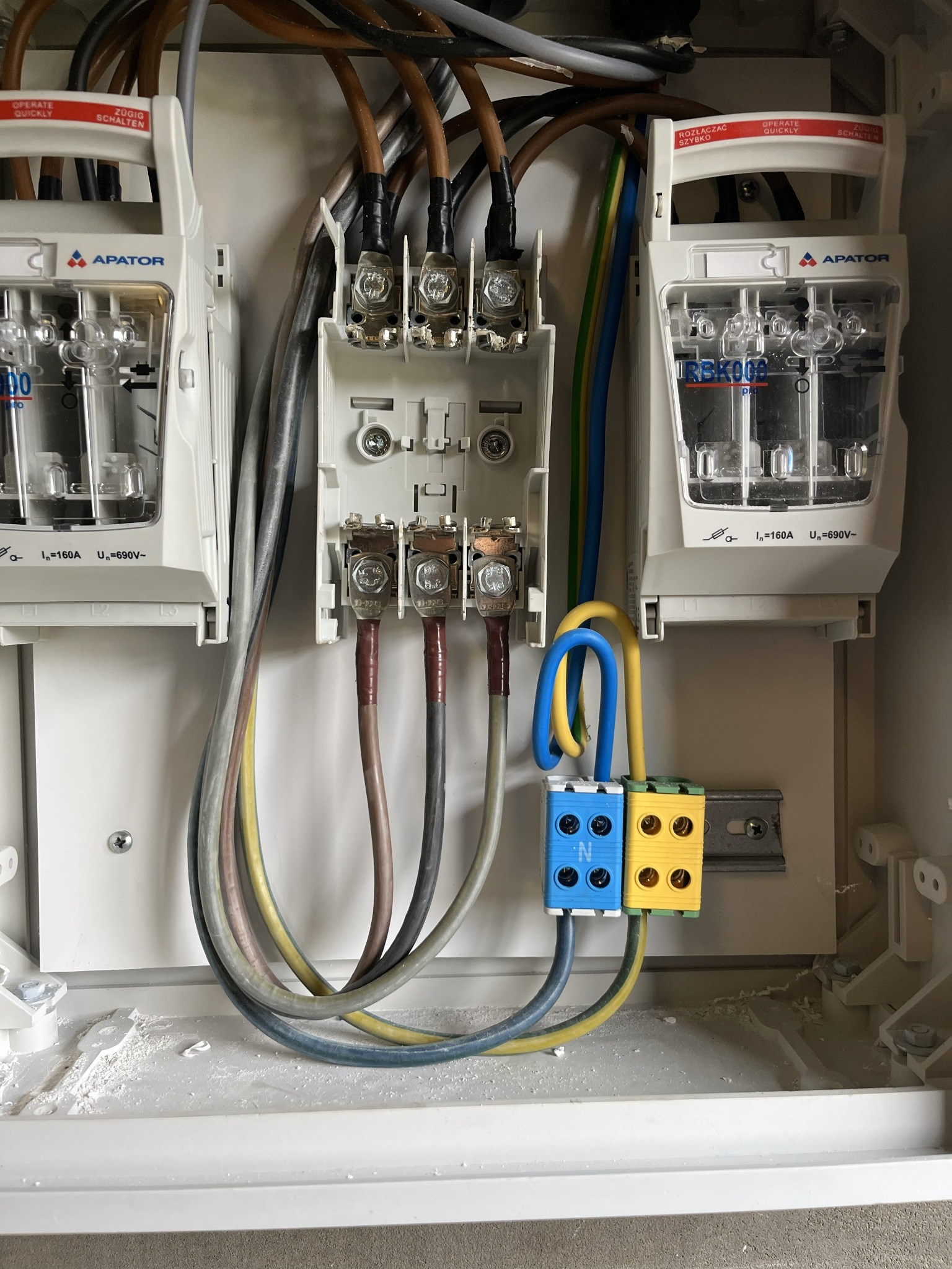 Szczegółowe ujęcie wnętrza skrzynki elektrycznej z bezpiecznikami Apator, widoczne przewody, złącza i elementy montażowe. Instalacja w trakcie prac, widoczne ślady po wierceniu.