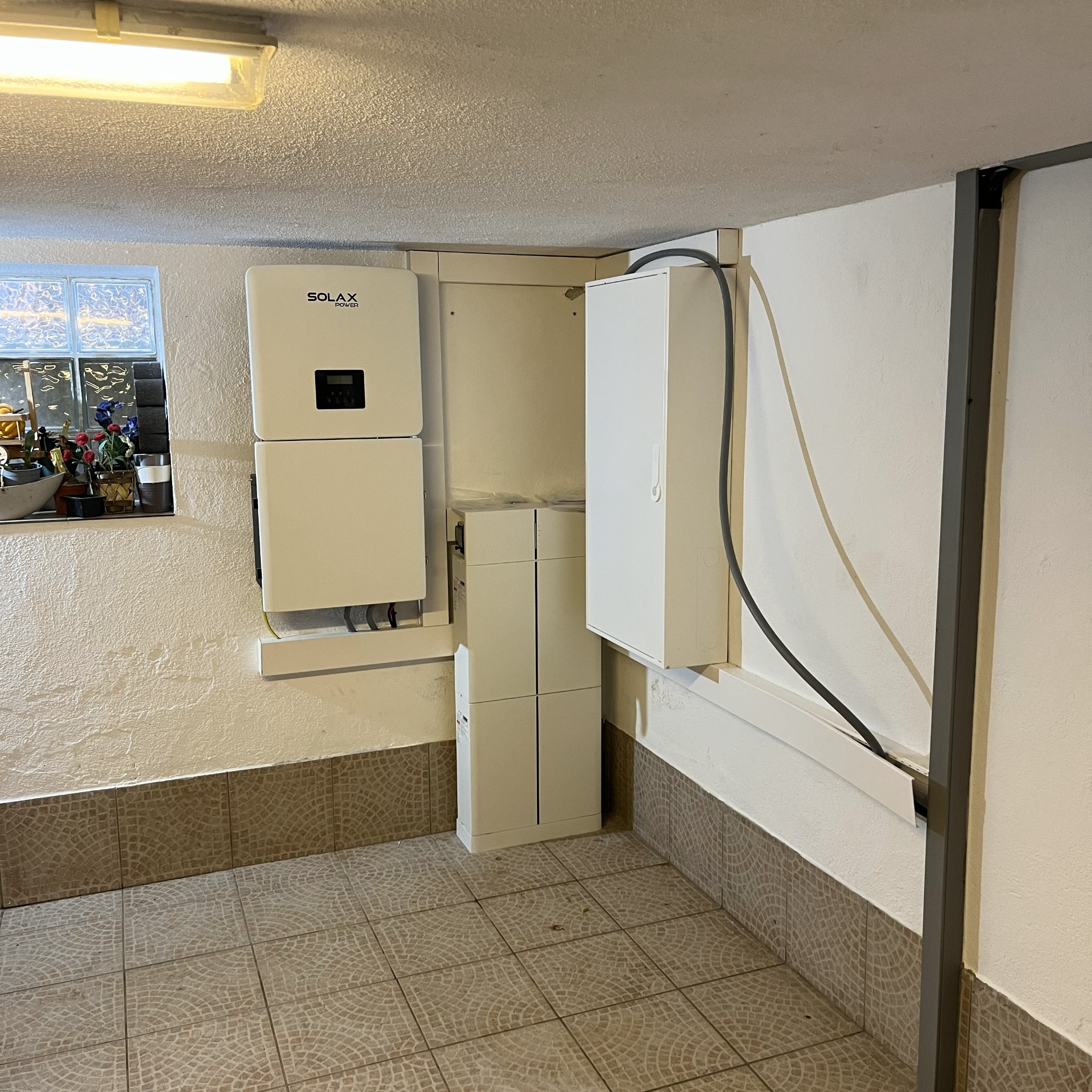 Biały inwerter Solax Power zamontowany na ścianie w pomieszczeniu z oknem i płytkami podłogowymi. Widoczne kable i dodatkowa skrzynka elektryczna.