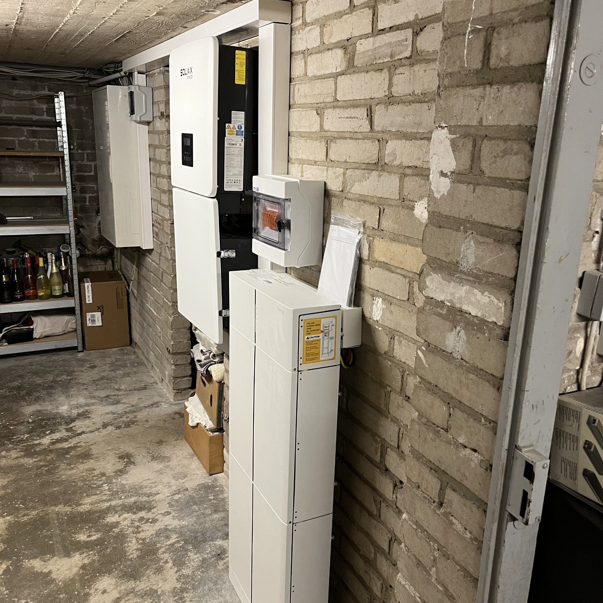 Biała szafa elektryczna Solax Power z rozdzielnicą na ceglanej ścianie w piwnicy. Widoczne kable i instalacje.