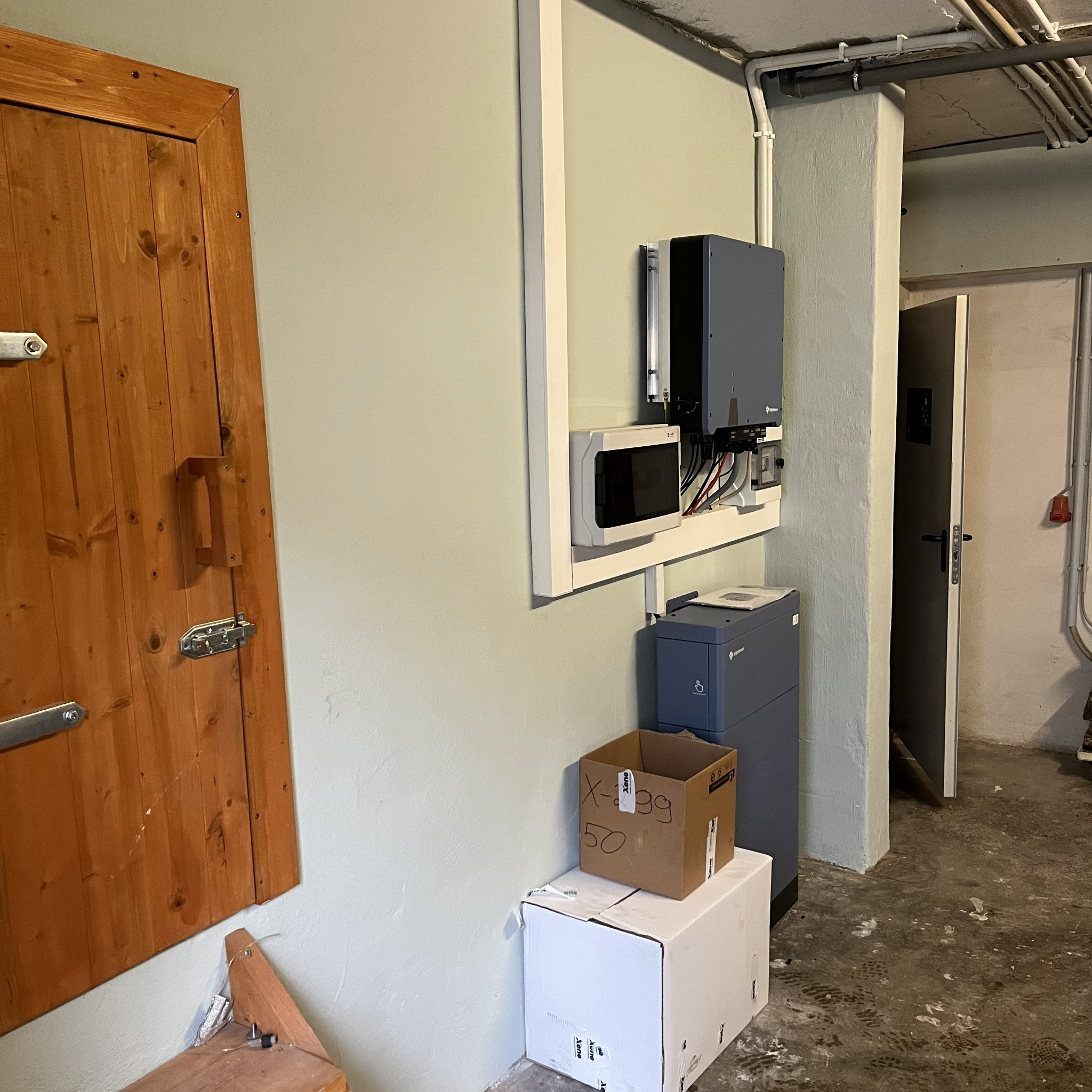 Szara szafka elektryczna z białym panelem sterowania zamontowana na ścianie w piwnicy, obok drewnianych drzwi i kartonów. Widoczne rury pod sufitem.