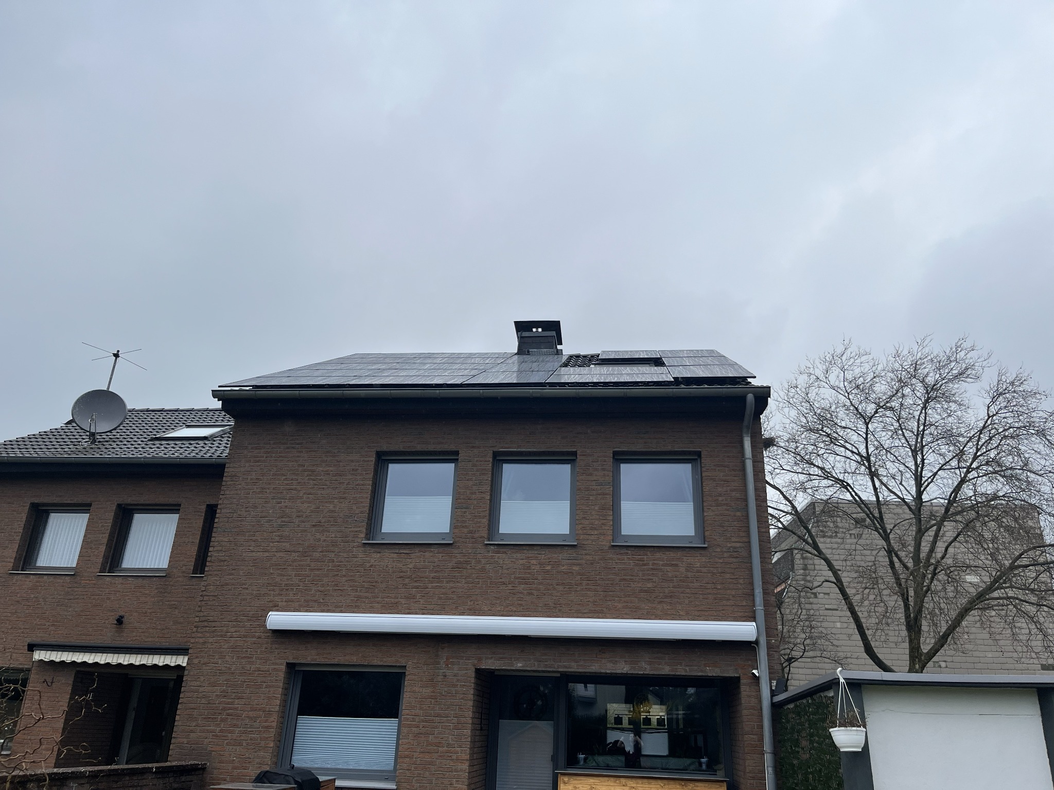 Brązowy dom z cegły z panelami słonecznymi na dachu w pochmurny dzień. Widoczna antena satelitarna i drzewo bez liści. Część paneli uszkodzona.