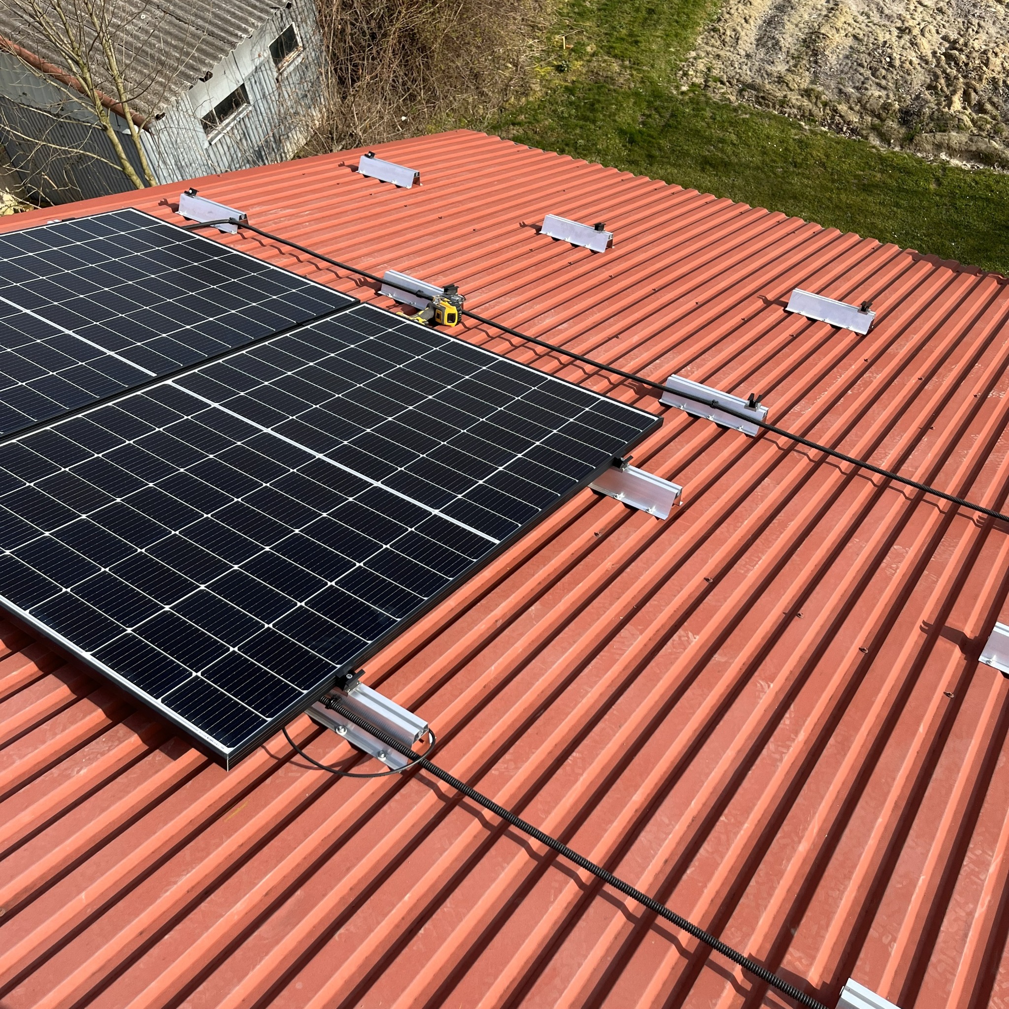Montaż paneli fotowoltaicznych na dachu z blachy w Rudzie Śląskiej. Widoczne panele, uchwyty montażowe i przewody. Dach w kolorze ceglastym, budynek w tle.
