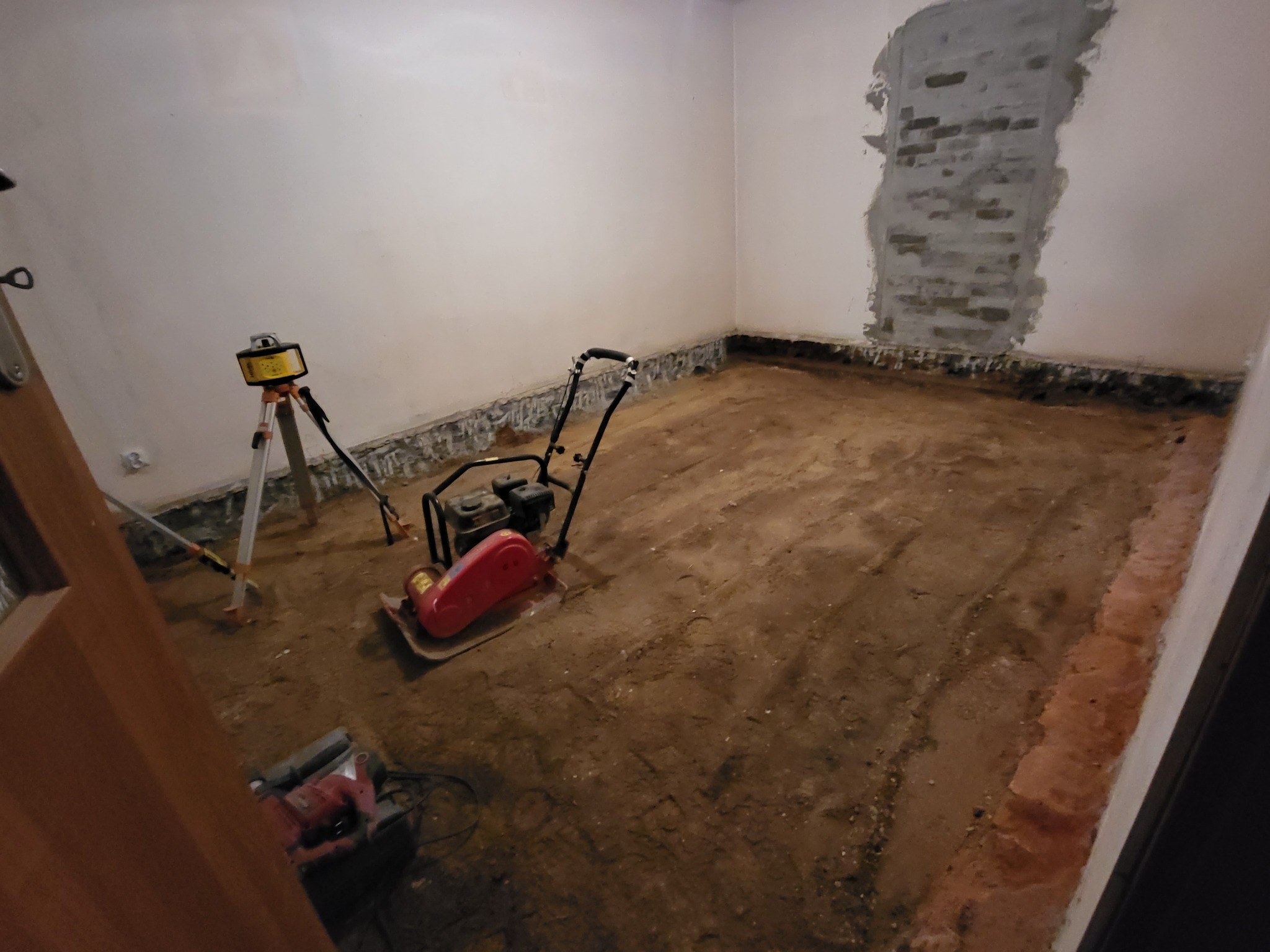 Przygotowanie podłoża: ubijarka w pomieszczeniu z odkrytą cegłą i niwelatorem laserowym. Etap prac przygotowawczych przed wylewką.