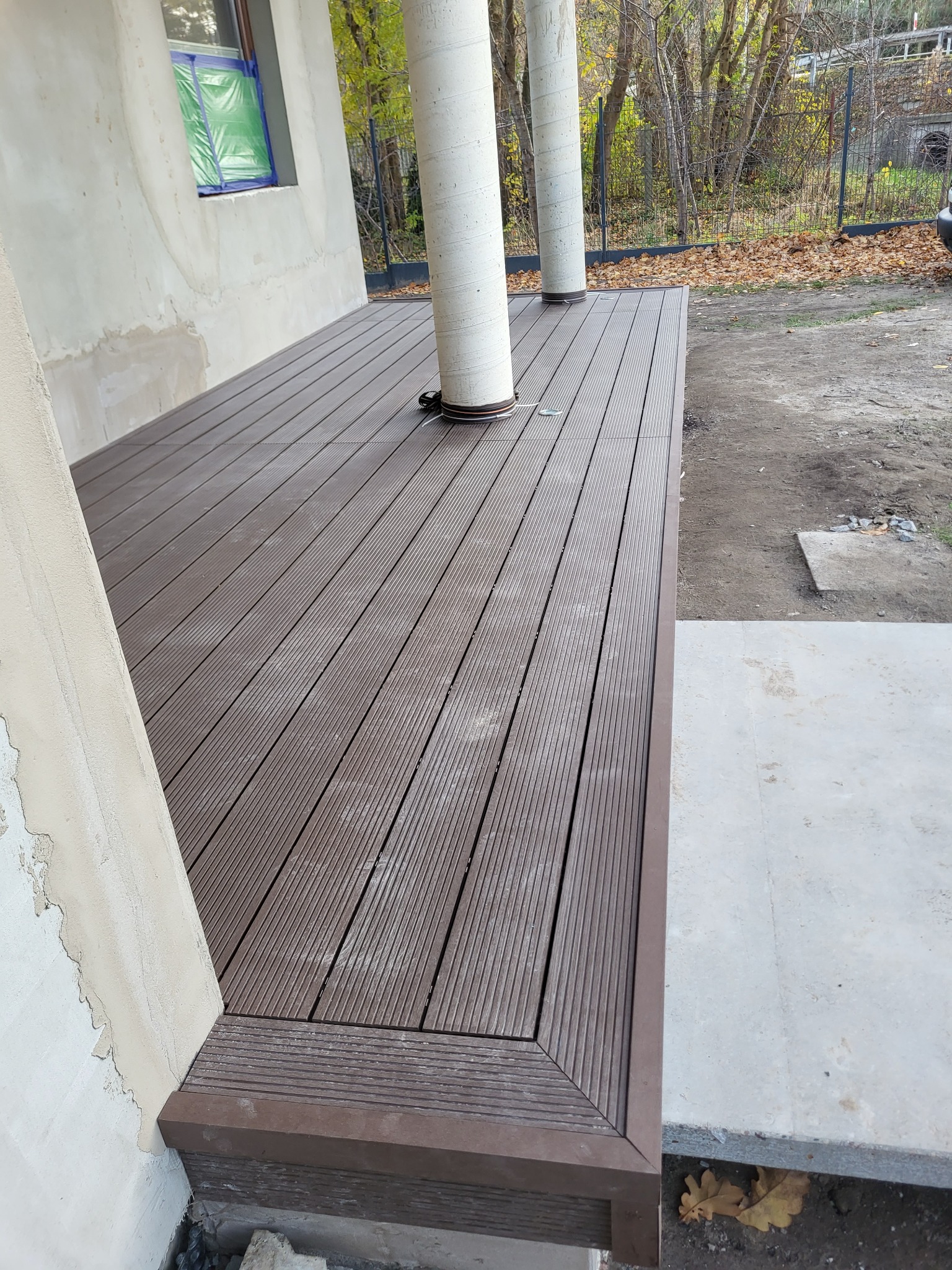 Nowy taras kompozytowy z brązowych desek, z widocznymi słupami i fragmentem elewacji w trakcie wykończenia. Jesienne otoczenie z opadłymi liśćmi.