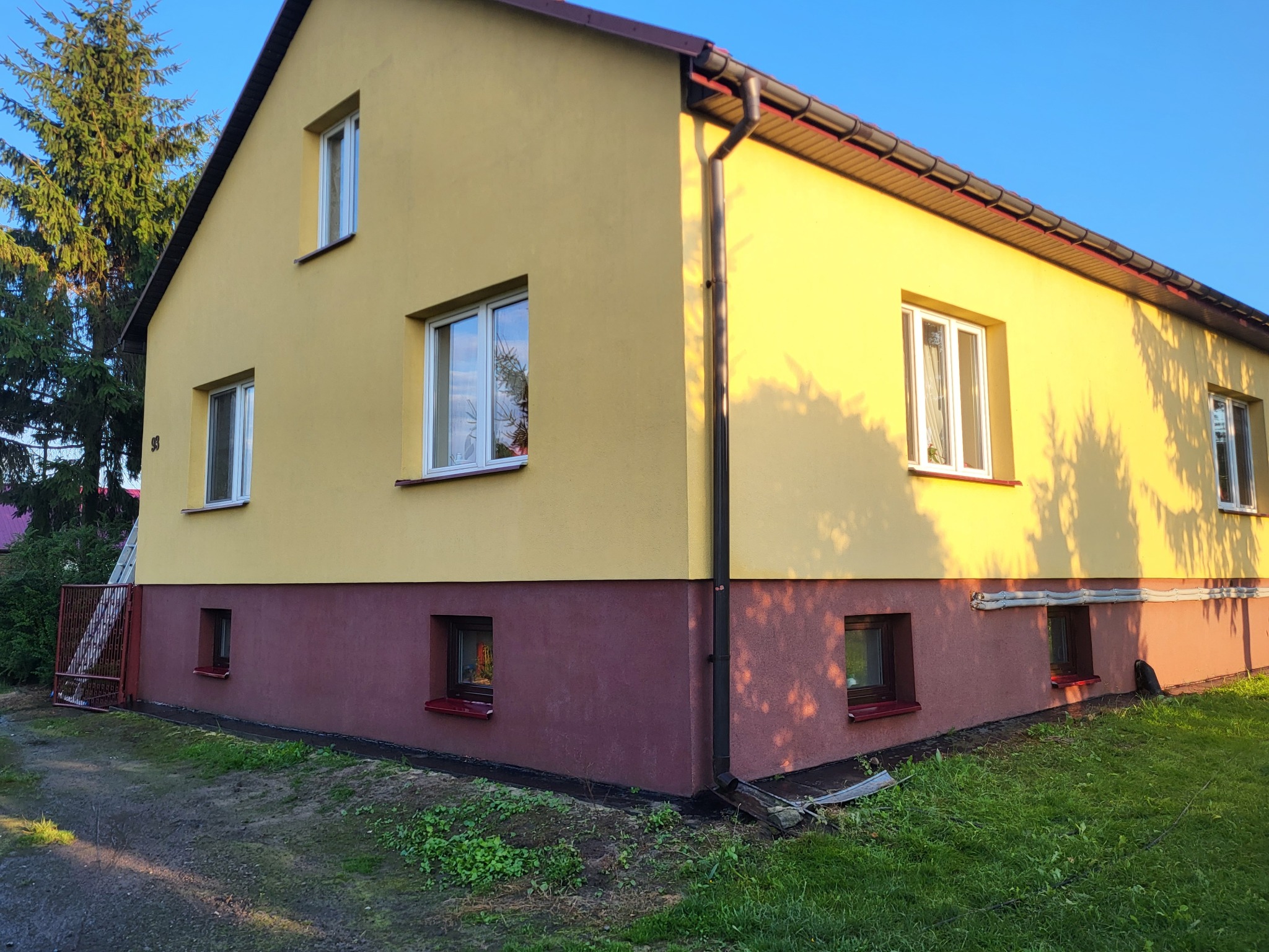 Dwukondygnacyjny dom z żółtą elewacją i bordową podmurówką, widoczny z poziomu gruntu, w słoneczny dzień. Widoczne okna z białymi ramami i brązowe rynny.