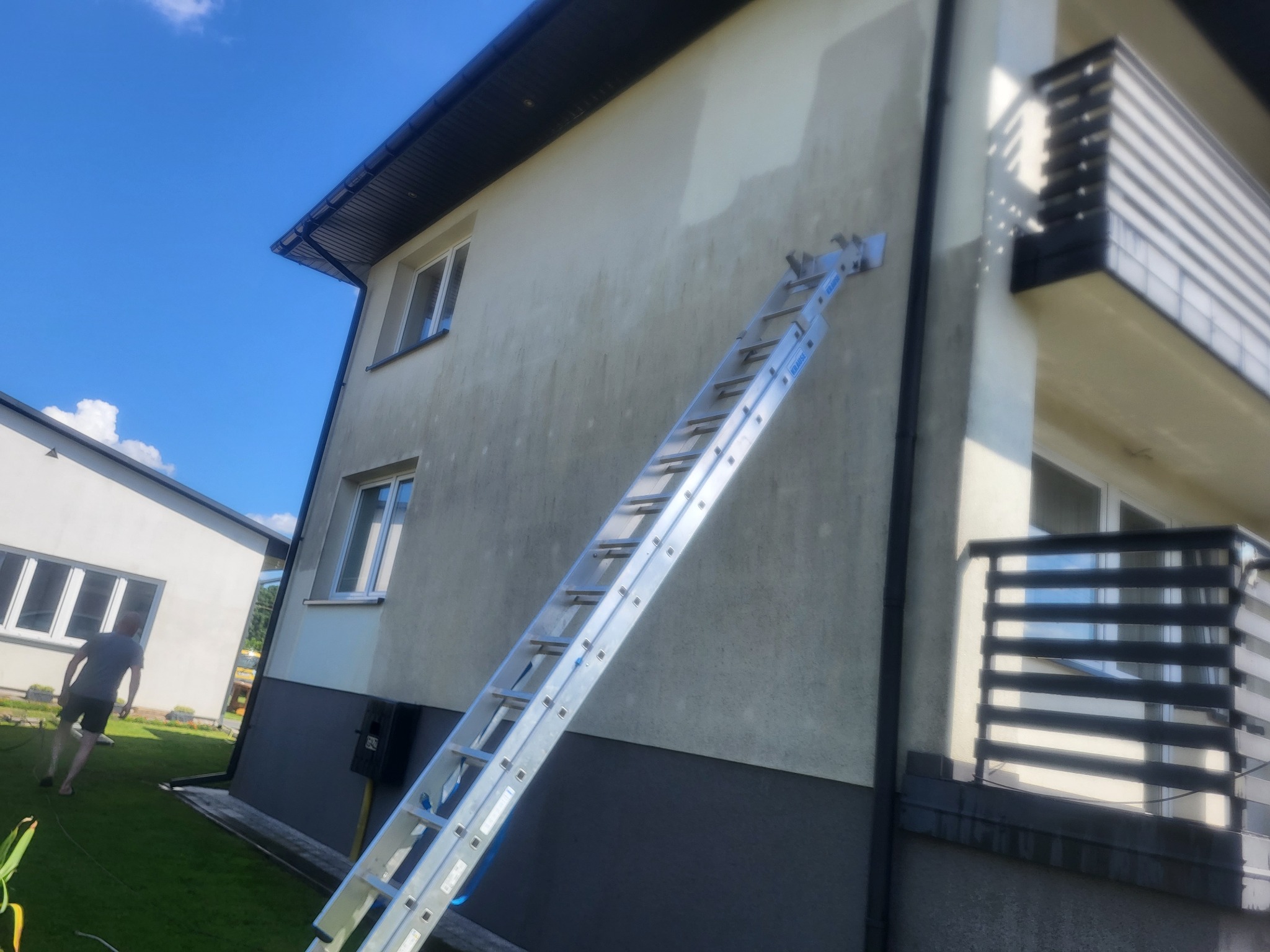 Dwupiętrowy dom z elewacją w trakcie czyszczenia. Aluminiowa drabina oparta o ścianę, widoczne różnice w czystości. Osoba w oddali na trawniku.