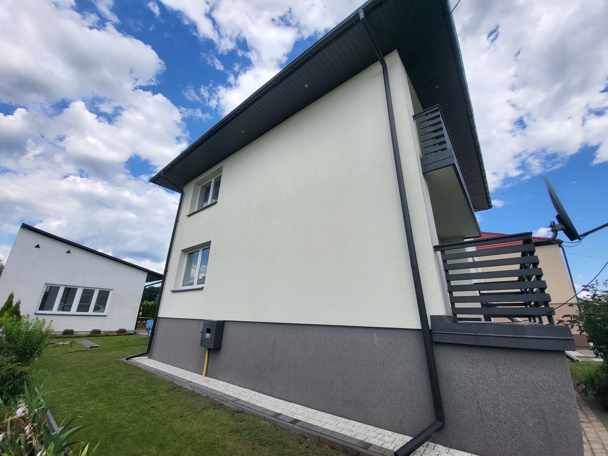 Elewacja domu z balkonem, świeżo po remoncie. Widoczne nowe okna, orynnowanie i wykończenie cokołu. Błękitne niebo z chmurami w tle.