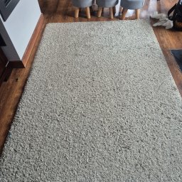 Fresh Tapicer Łukasz Miller - Jasnoszary dywan shaggy na drewnianej podłodze, widoczne trzy szare stołki i fragment węża odkurzacza. Tekstura dywanu wyraźnie widoczna.