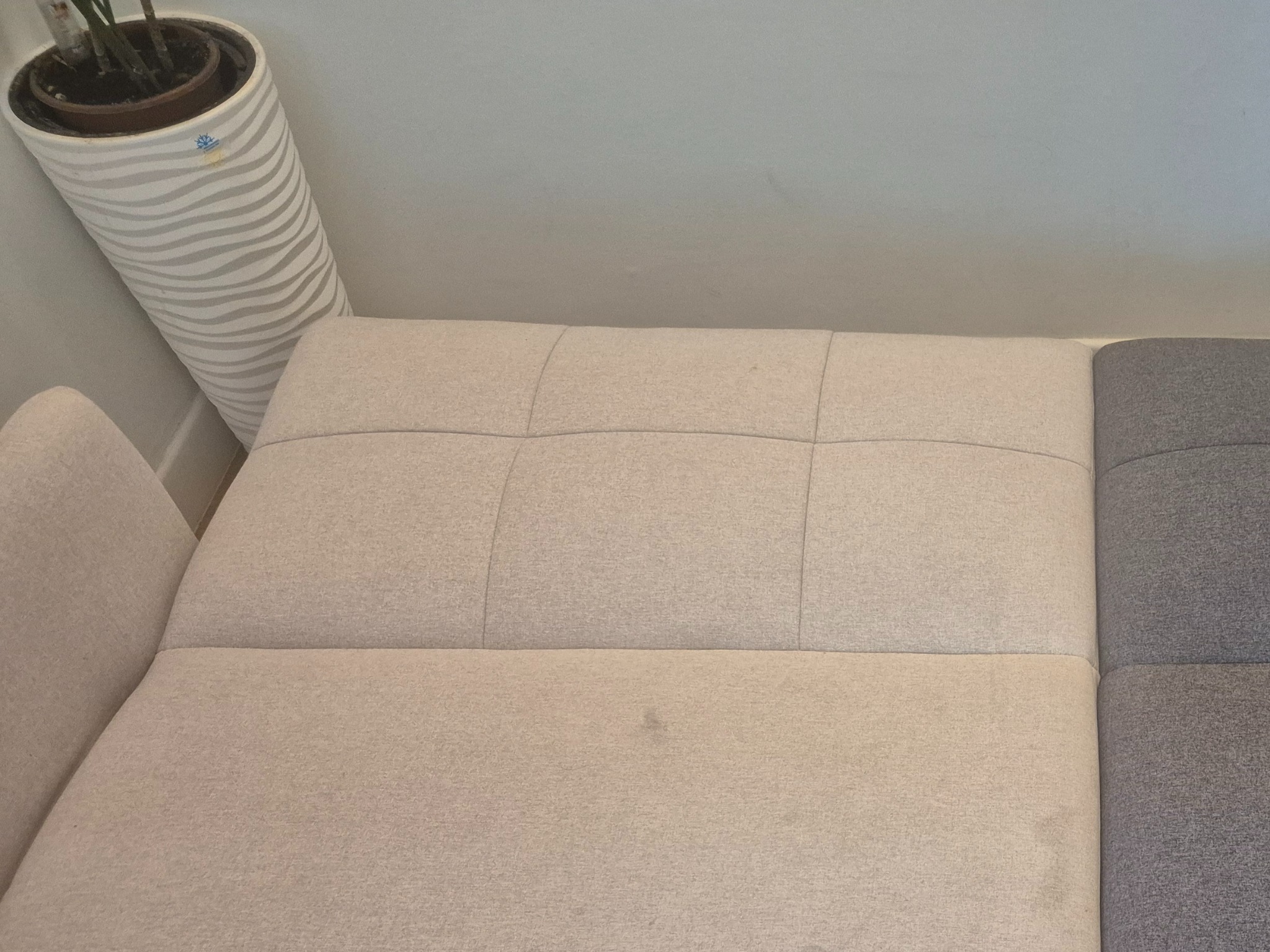 Beżowa kanapa z plamą, obok szara część i wysoka donica w paski. Widok z góry, naturalne światło. Wymaga prania tapicerki.