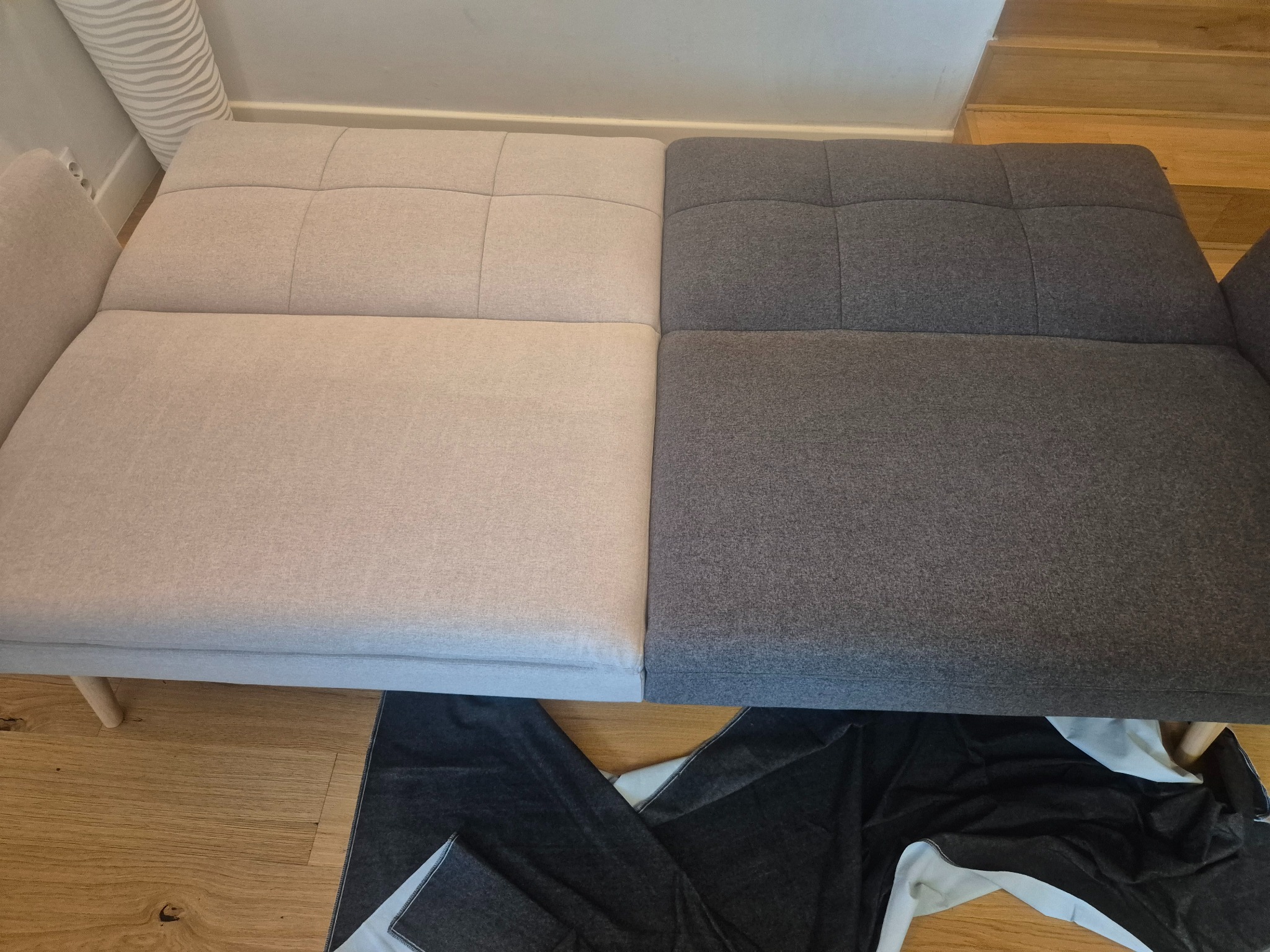 Rozłożona kanapa z dwukolorową tapicerką (beż i szary) na drewnianej podłodze, widoczny fragment czarnego materiału pod kanapą. Widok z góry.