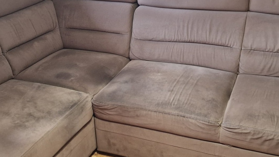 Szara, narożna kanapa z widocznymi plamami na siedziskach, wymagająca odświeżenia. Widoczne ślady użytkowania na tapicerce.