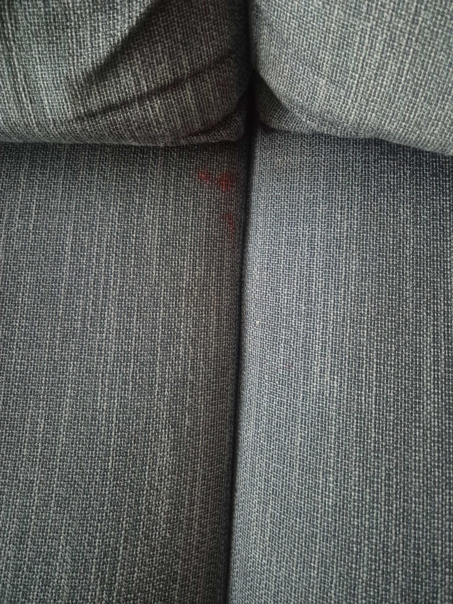 Szary materiał tapicerki kanapy z widocznym, czerwonym zabrudzeniem między siedziskami. Tekstura tkaniny wyraźnie widoczna.