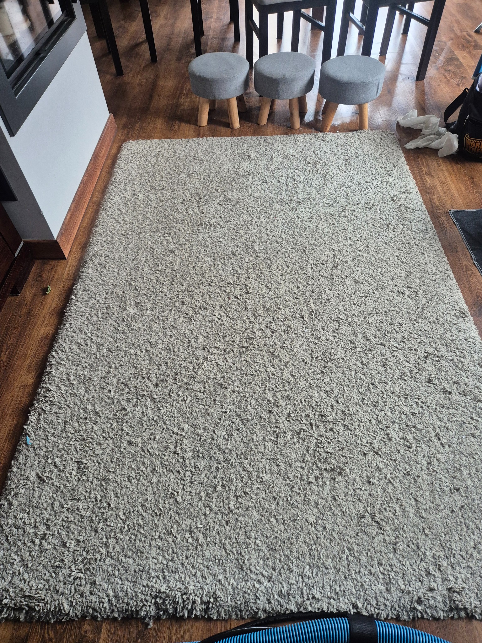 Jasnoszary dywan shaggy na drewnianej podłodze, widoczne trzy szare stołki i fragment węża odkurzacza. Tekstura dywanu wyraźnie widoczna.