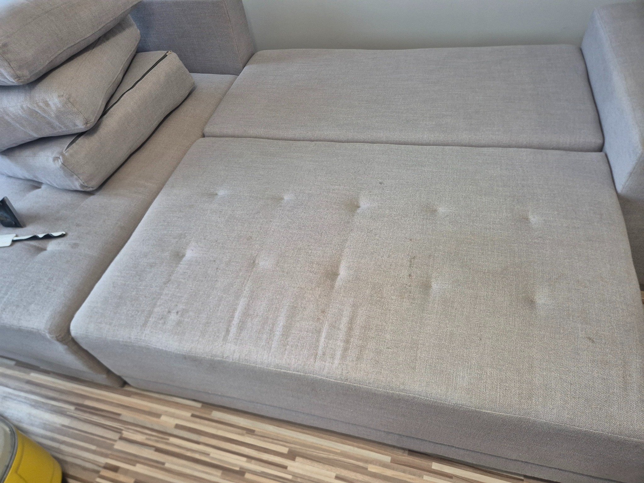 Jasnoszara, modułowa kanapa z widocznymi plamami na siedzisku, sugerująca potrzebę czyszczenia tapicerki. Częściowo widoczny odkurzacz piorący.