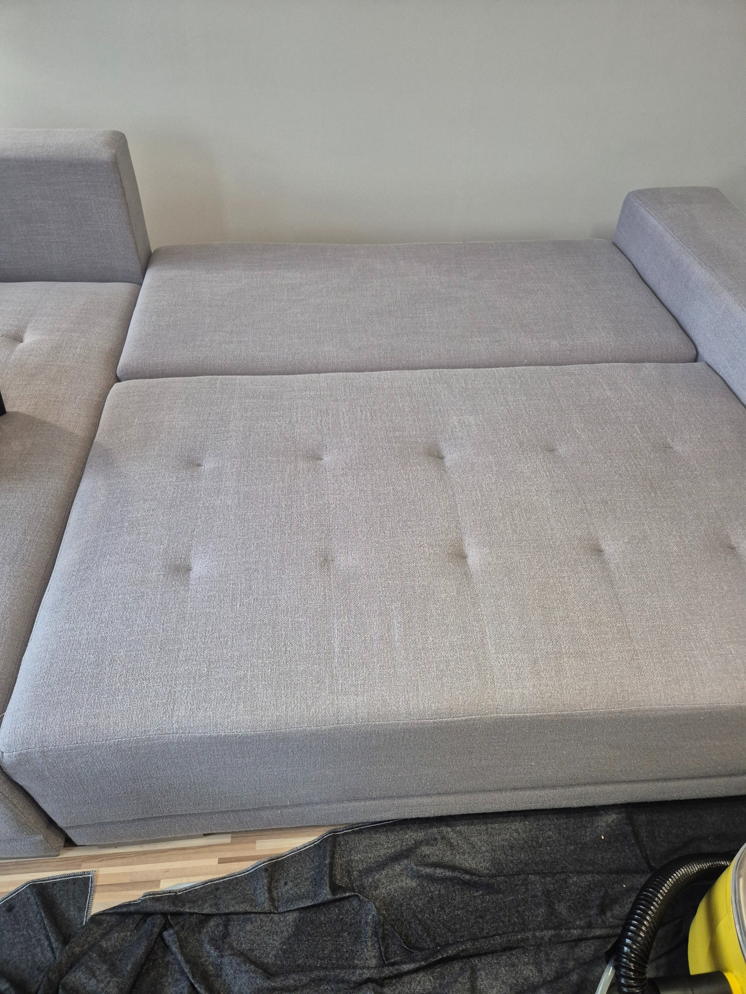 Jasnoszara, modułowa kanapa z tapicerowanymi guzikami, częściowo przykryta czarną folią ochronną, z widocznym fragmentem węża odkurzacza piorącego.
