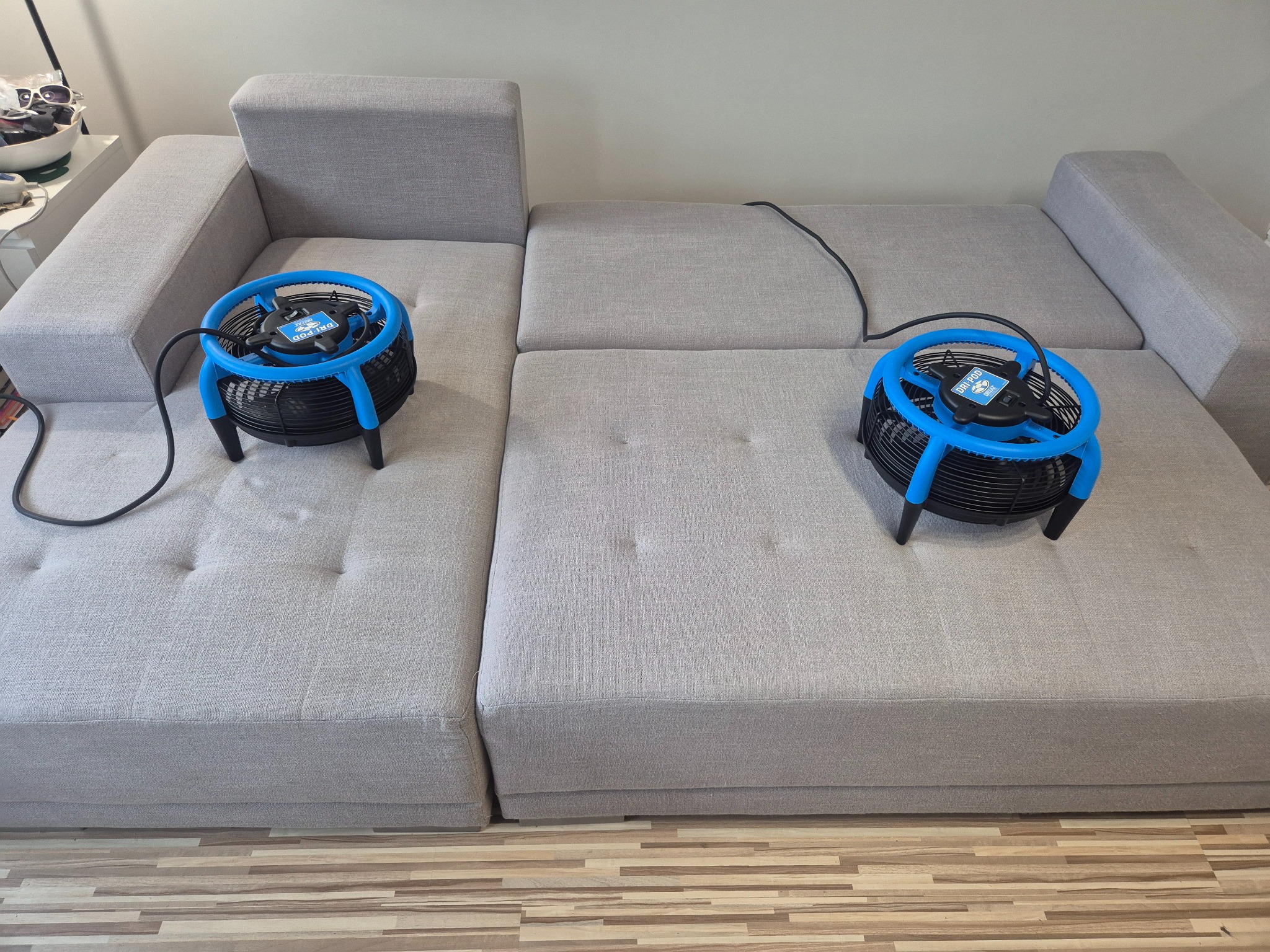 Jasnoszara sofa z dwoma niebieskimi wentylatorami do suszenia tapicerki po praniu, ustawionymi na siedziskach. Widok z góry, parkiet na podłodze.