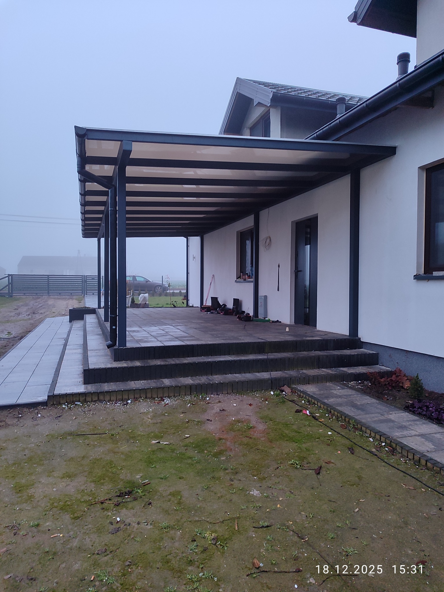 Nowoczesna, grafitowa pergola przy białym domu w pochmurny dzień. Widoczne schody z kostki brukowej i fragment ogrodu. Minimalistyczny design.