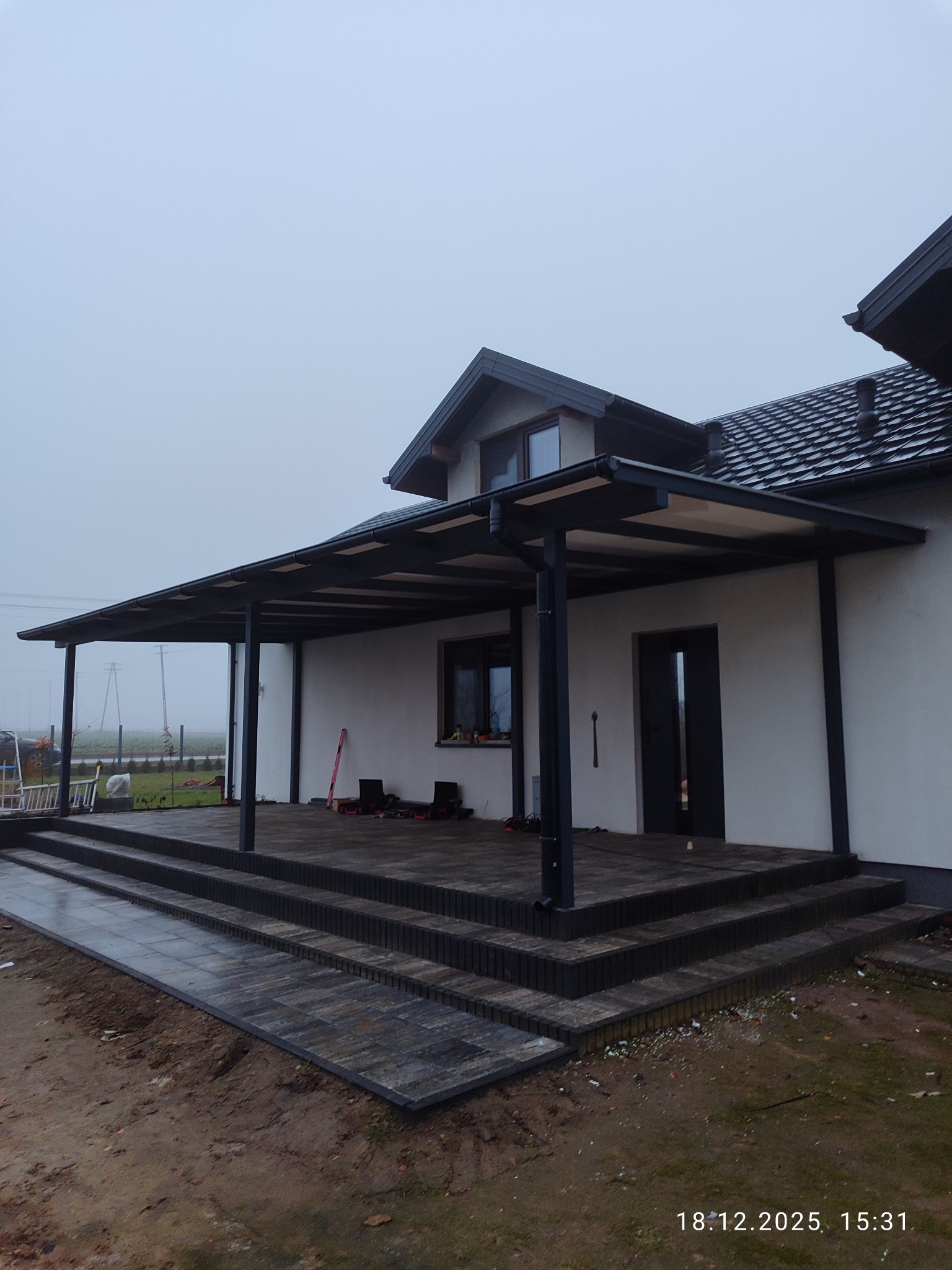 Nowoczesna, grafitowa pergola metalowa przy białym domu z brukowanym tarasem i schodami. Widok na zewnątrz w pochmurny dzień. Minimalistyczny design.