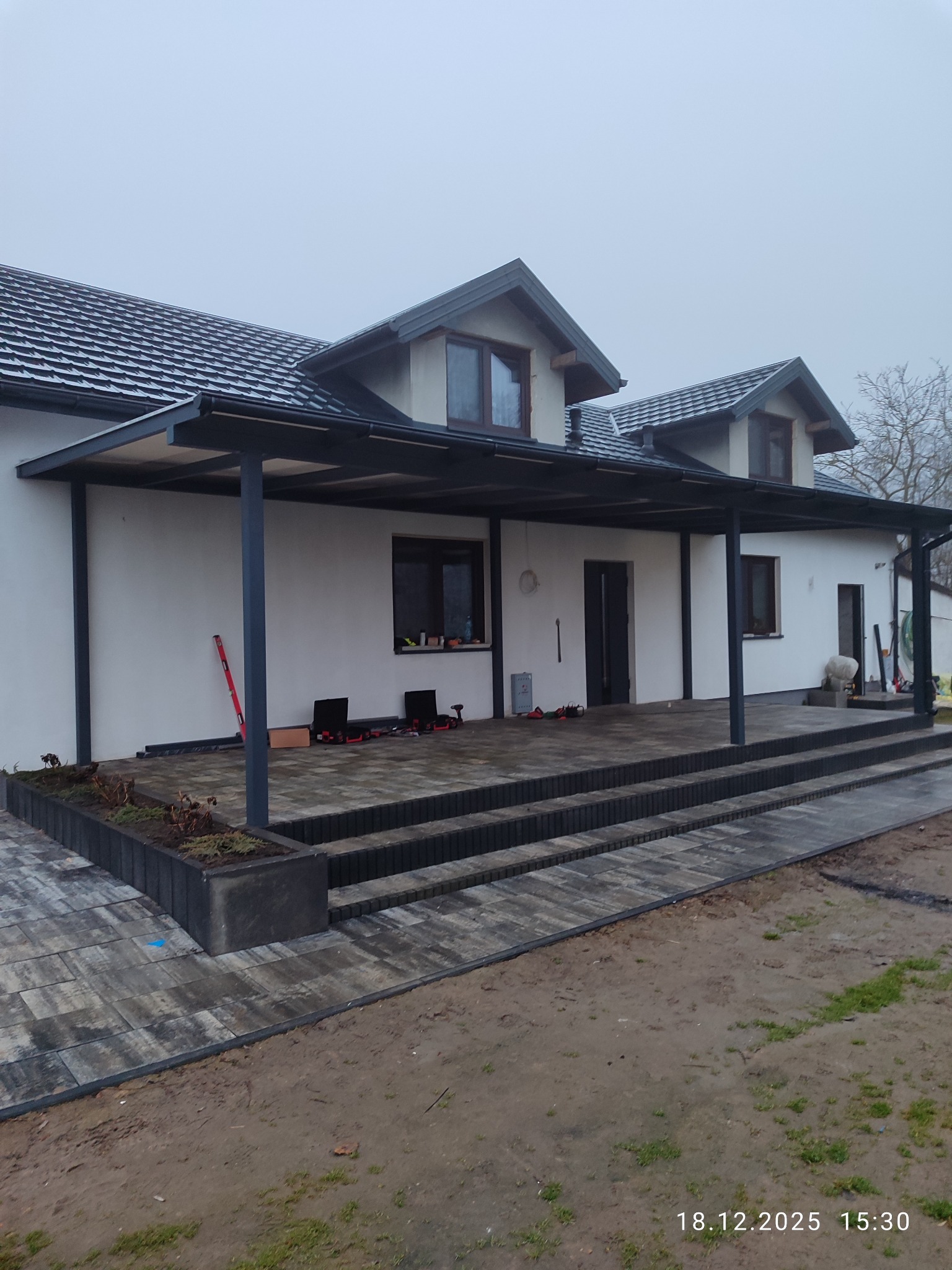 Nowoczesna, grafitowa pergola metalowa przy białym domu z kostką brukową. Widoczne narzędzia i poziomica, sugerujące etap montażu. Pochmurne niebo w tle.