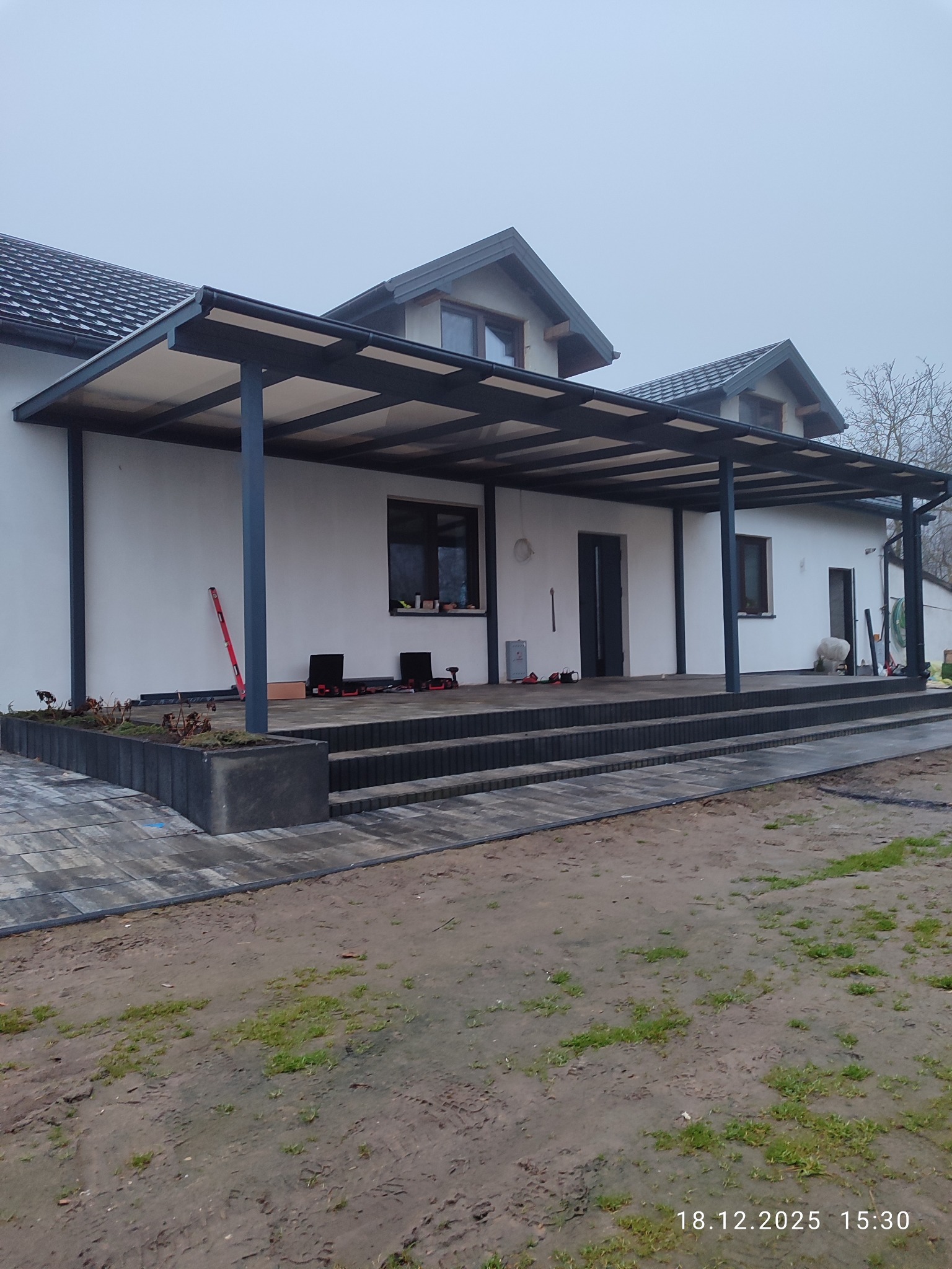 Nowoczesna, metalowa pergola przy białym domu z ciemnymi oknami. Widoczne narzędzia i poziomica sugerują etap prac wykończeniowych. Uwagę przyciąga minimalistyczny design.