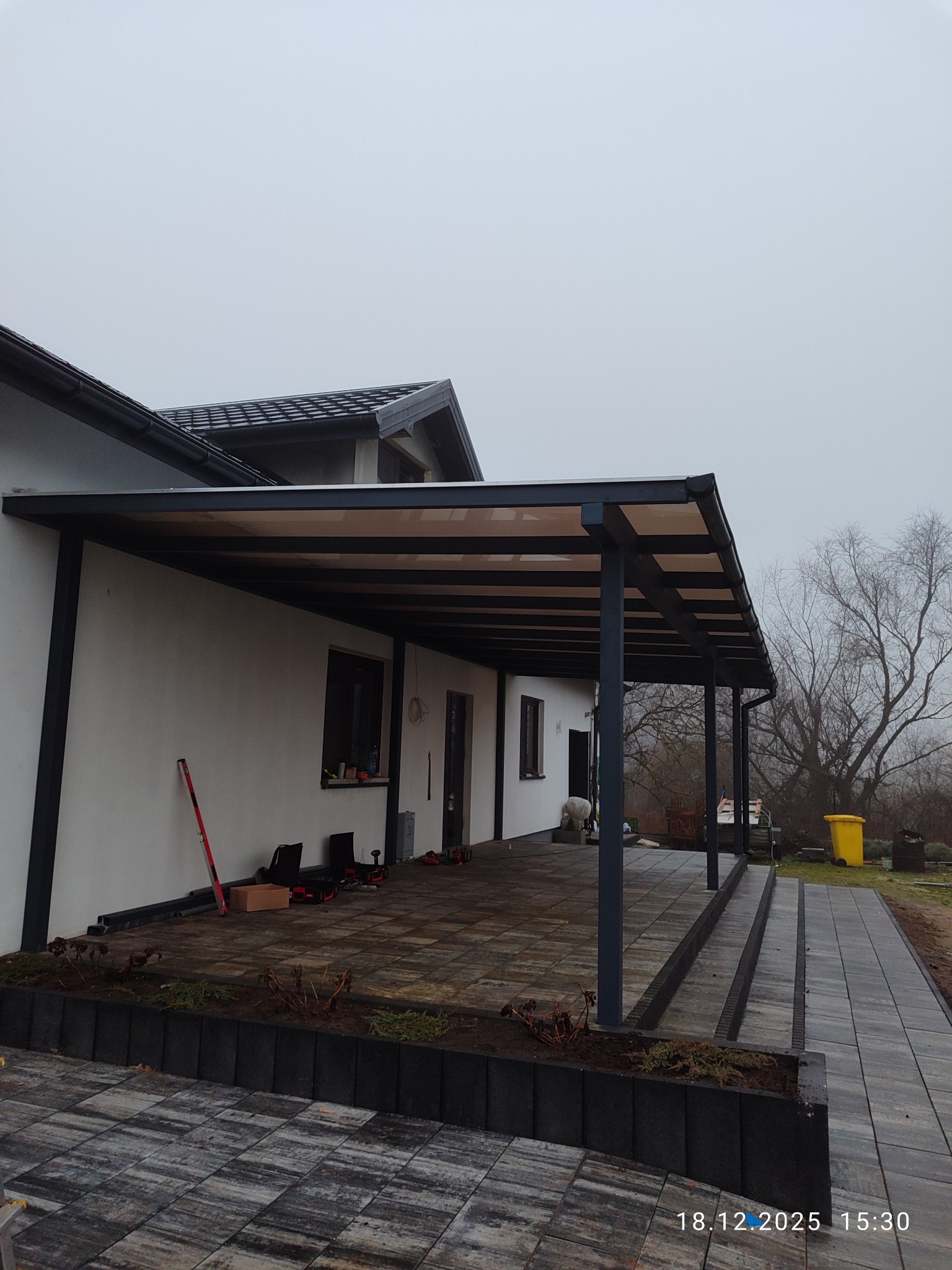 Nowoczesna, grafitowa pergola metalowa przy białym domu z ciemną dachówką. Widoczne narzędzia i kostka brukowa. Pochmurny dzień.