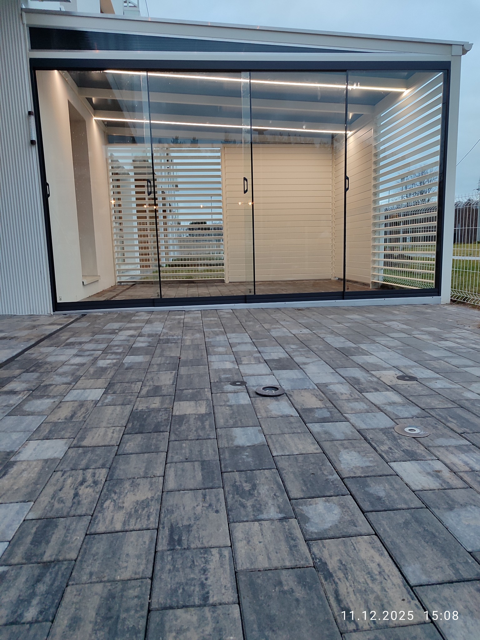 Nowoczesna zabudowa balkonu z przesuwnymi szklanymi drzwiami i oświetleniem LED, widok z zewnątrz na kostkę brukową. Minimalistyczny design.