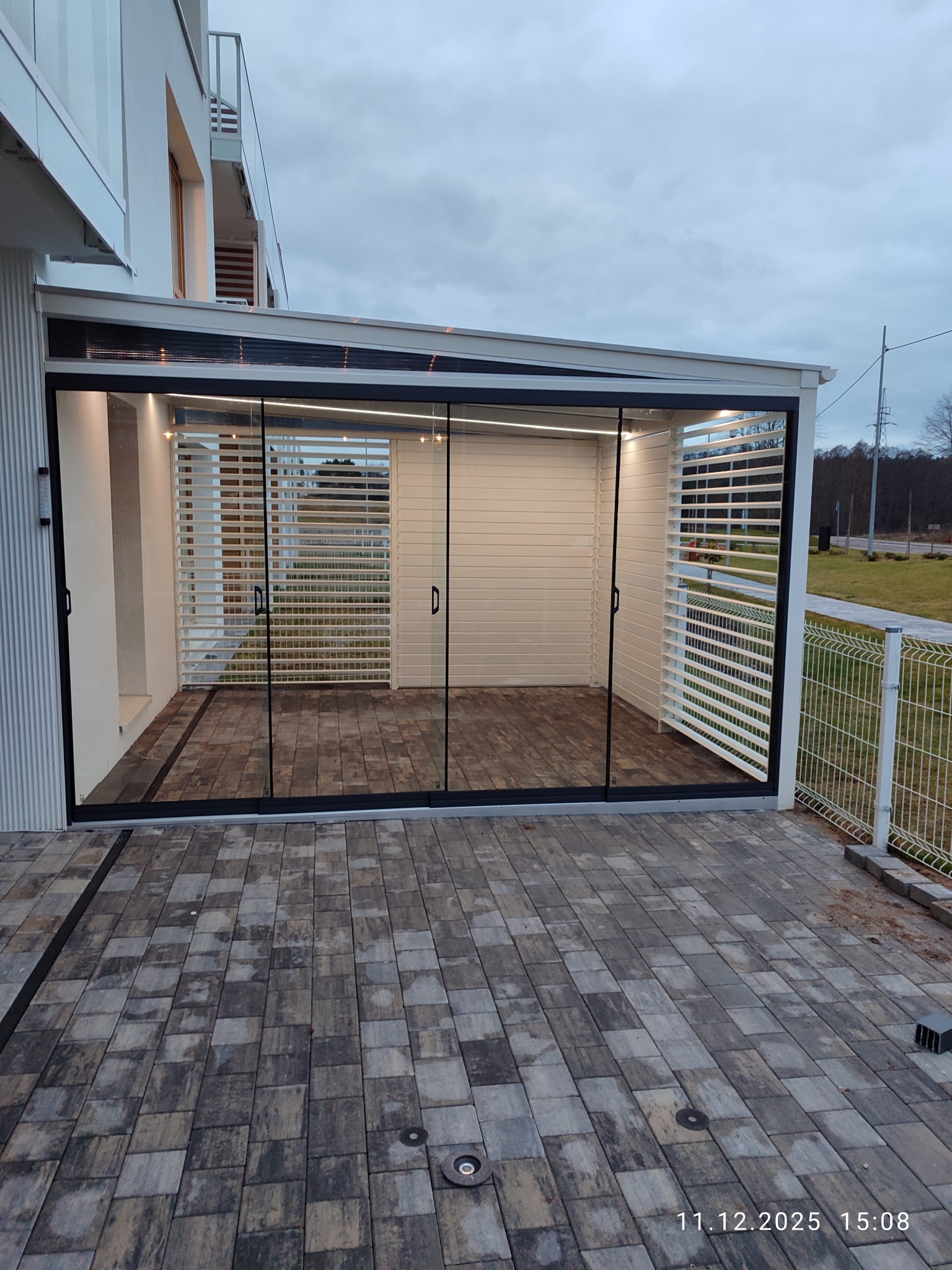 Nowoczesna zabudowa balkonu z przesuwnymi szklanymi drzwiami i białymi żaluzjami, oświetlona listwami LED, z widokiem na kostkę brukową i ogrodzenie.