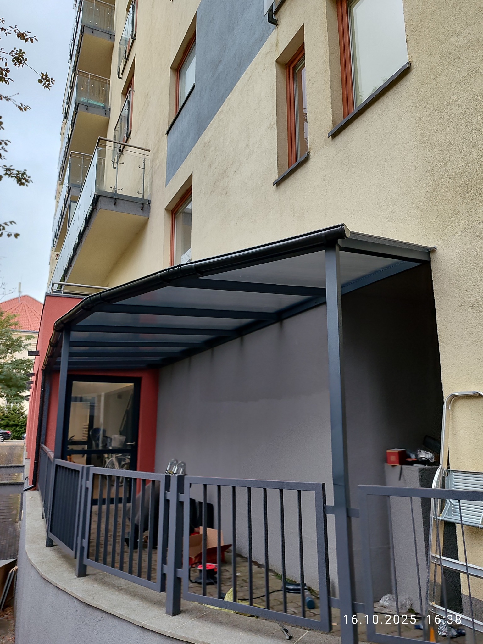 Metalowa pergola przy budynku mieszkalnym z balkonami, widok z dołu. Szara konstrukcja z przezroczystym zadaszeniem. Częściowo widoczne narzędzia i akcesoria montażowe.